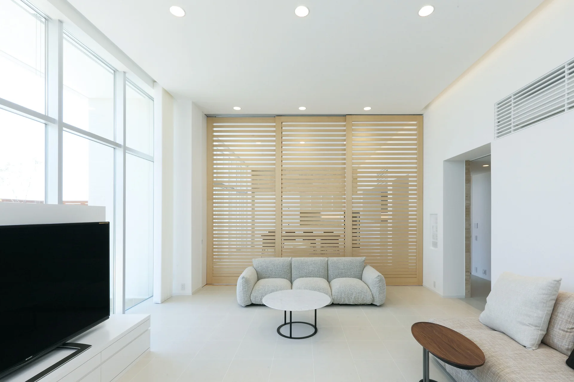 A minimalist living room featuring a light grey low-profile sofa, a round marble-topped coffee table, and a large slatted wood sliding screen.
