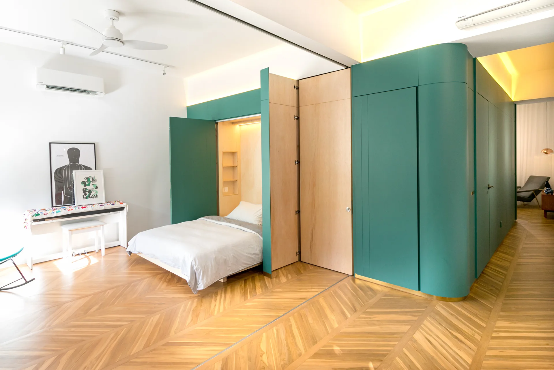 A bed is recessed within a custom teal and natural wood cabinet unit on light herringbone patterned wood floors.