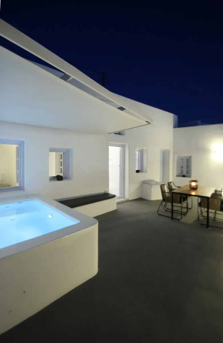 A minimalist outdoor terrace at night featuring a built-in white plaster hot tub, dark grey floor, and a small dining set under a dark blue sky.