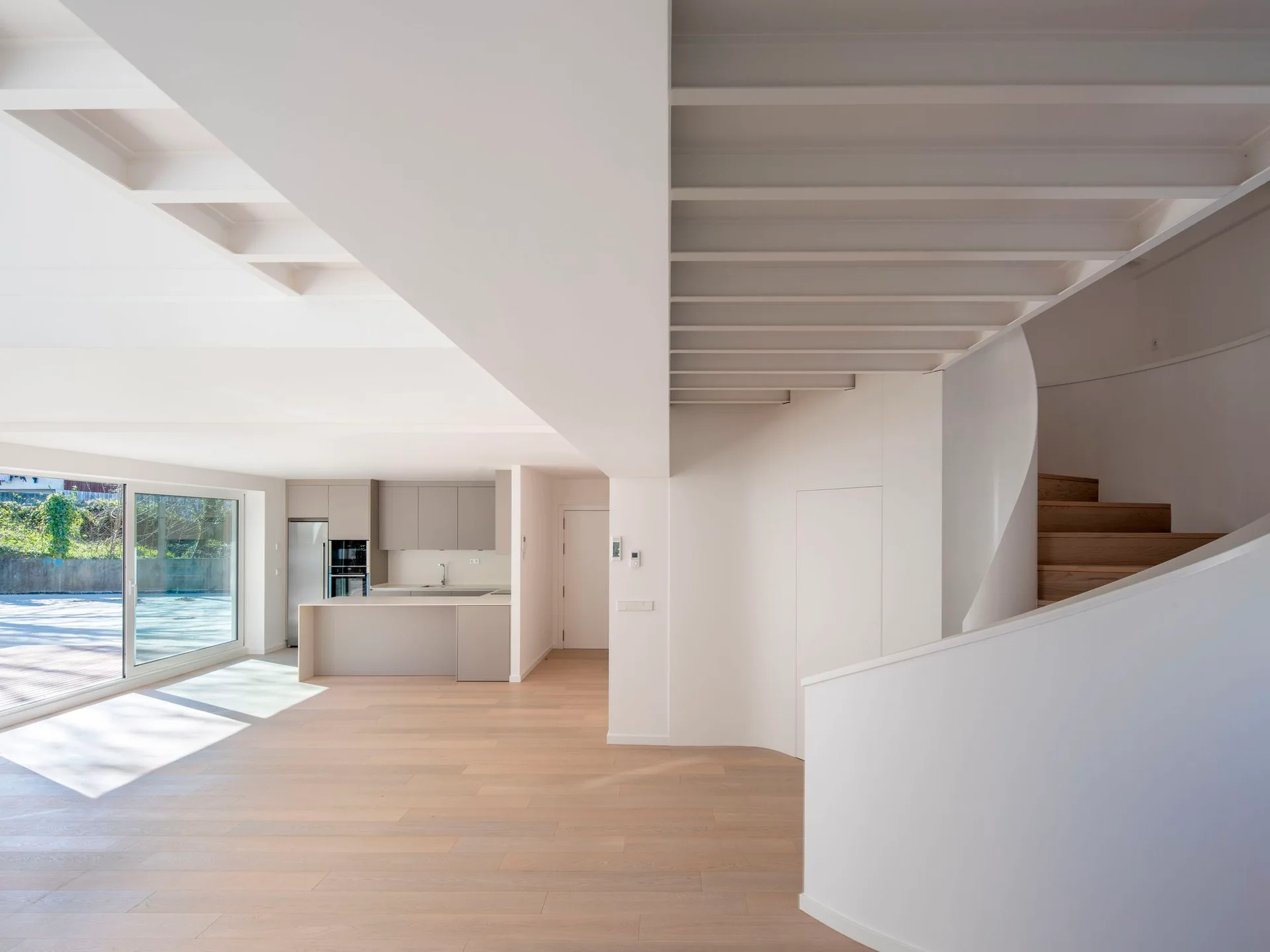 A minimalist white living area features a curved sculptural staircase with wood treads, light wood floors, and an open kitchen.