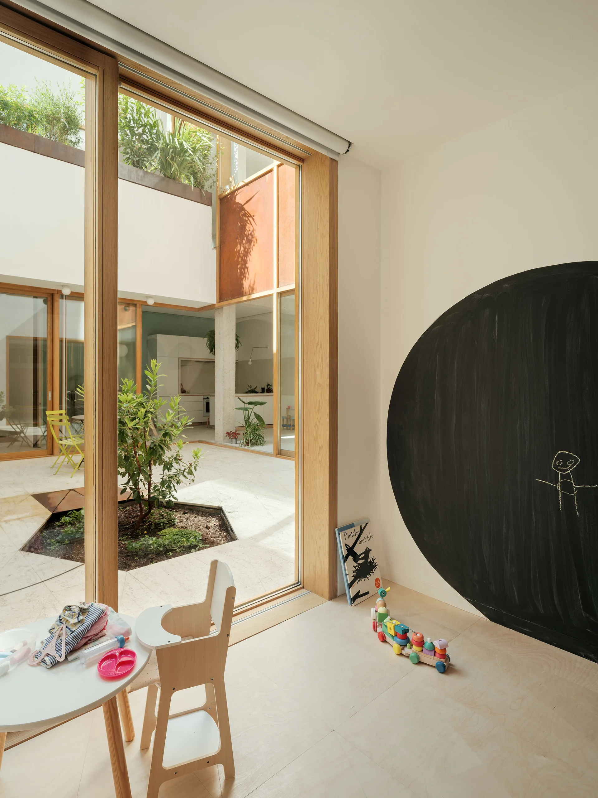 A bright children's play area with light wood floors features a large circular chalkboard, a small wooden high chair, and a sliding glass door to a courtyard.