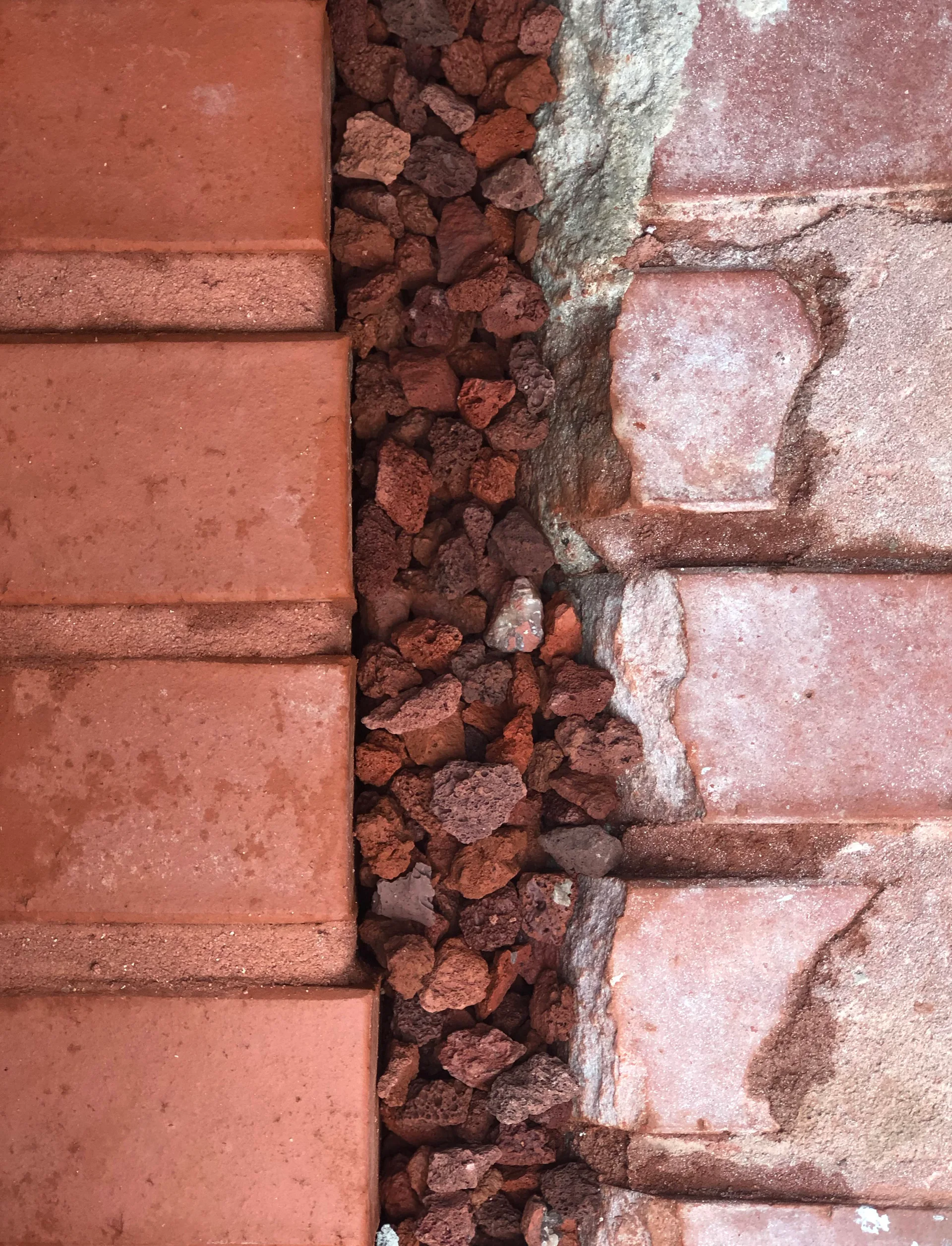 A top-down view of smooth red clay brick pavers separated from weathered masonry blocks by a narrow border filled with porous red volcanic gravel.