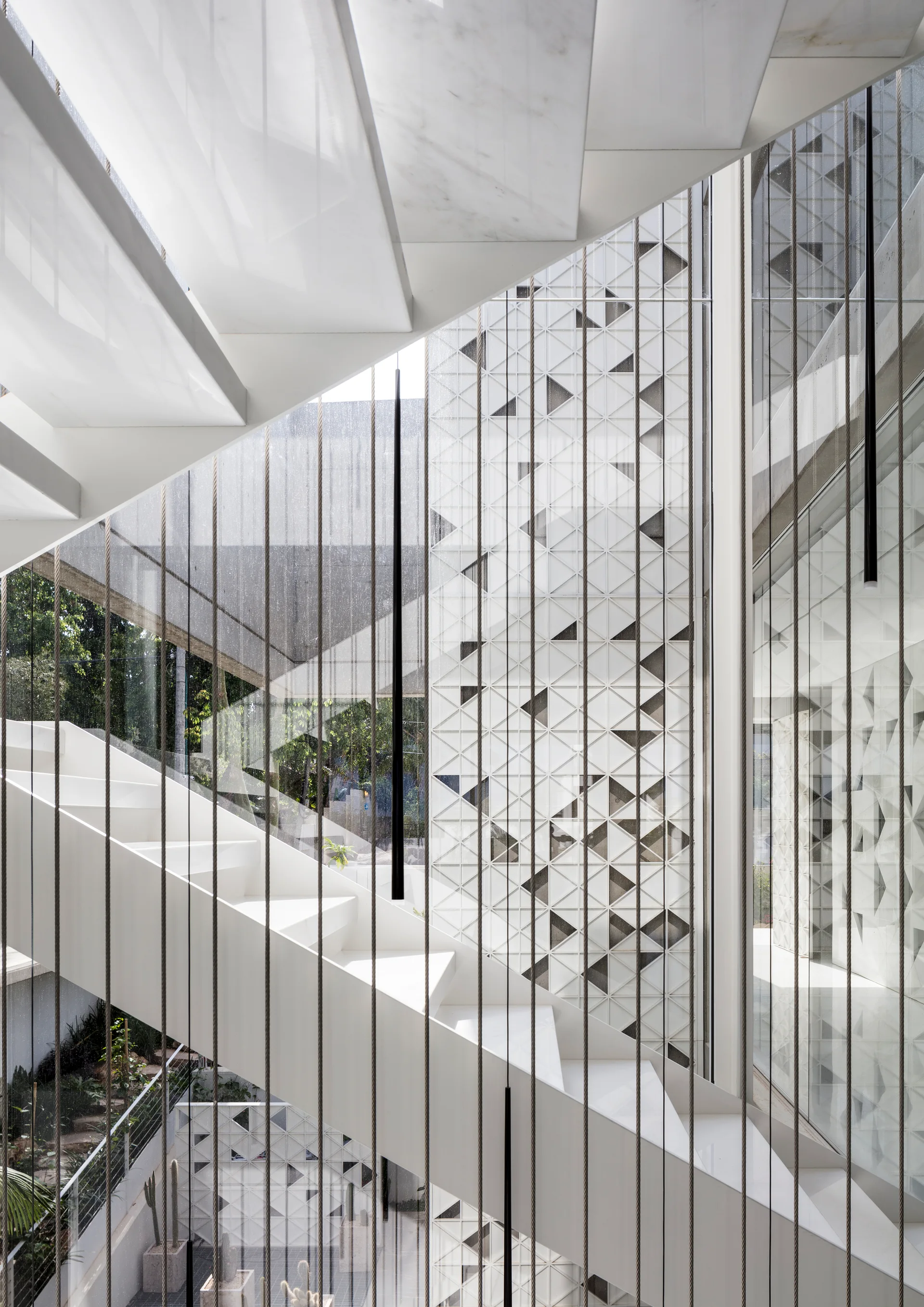 A modern white floating staircase is framed by thin vertical tension cables, a glass facade, and a geometric patterned wall.