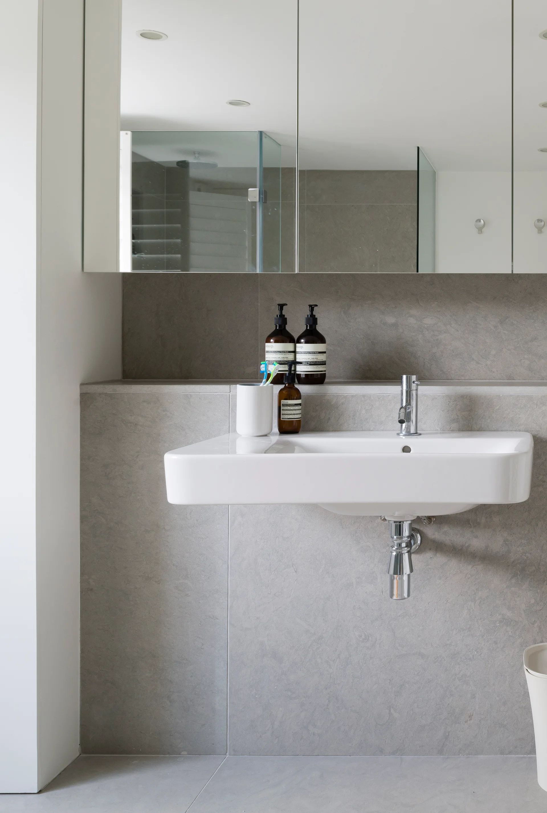 A white wall-mounted sink with a chrome faucet is set against large gray stone tiles under a mirrored medicine cabinet.