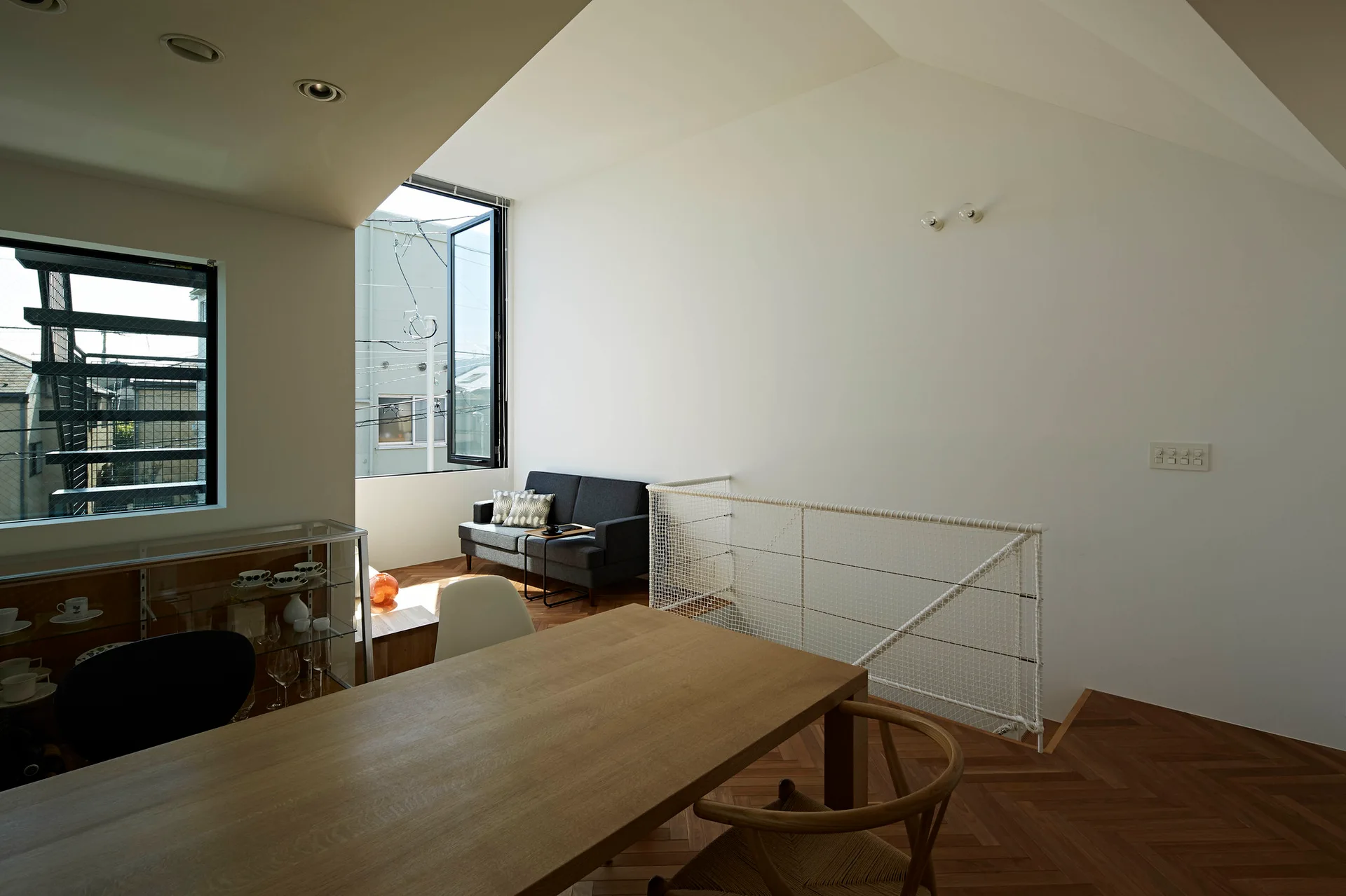 A wooden dining table sits on a herringbone patterned wood floor next to a white mesh staircase railing and a sloped white ceiling.