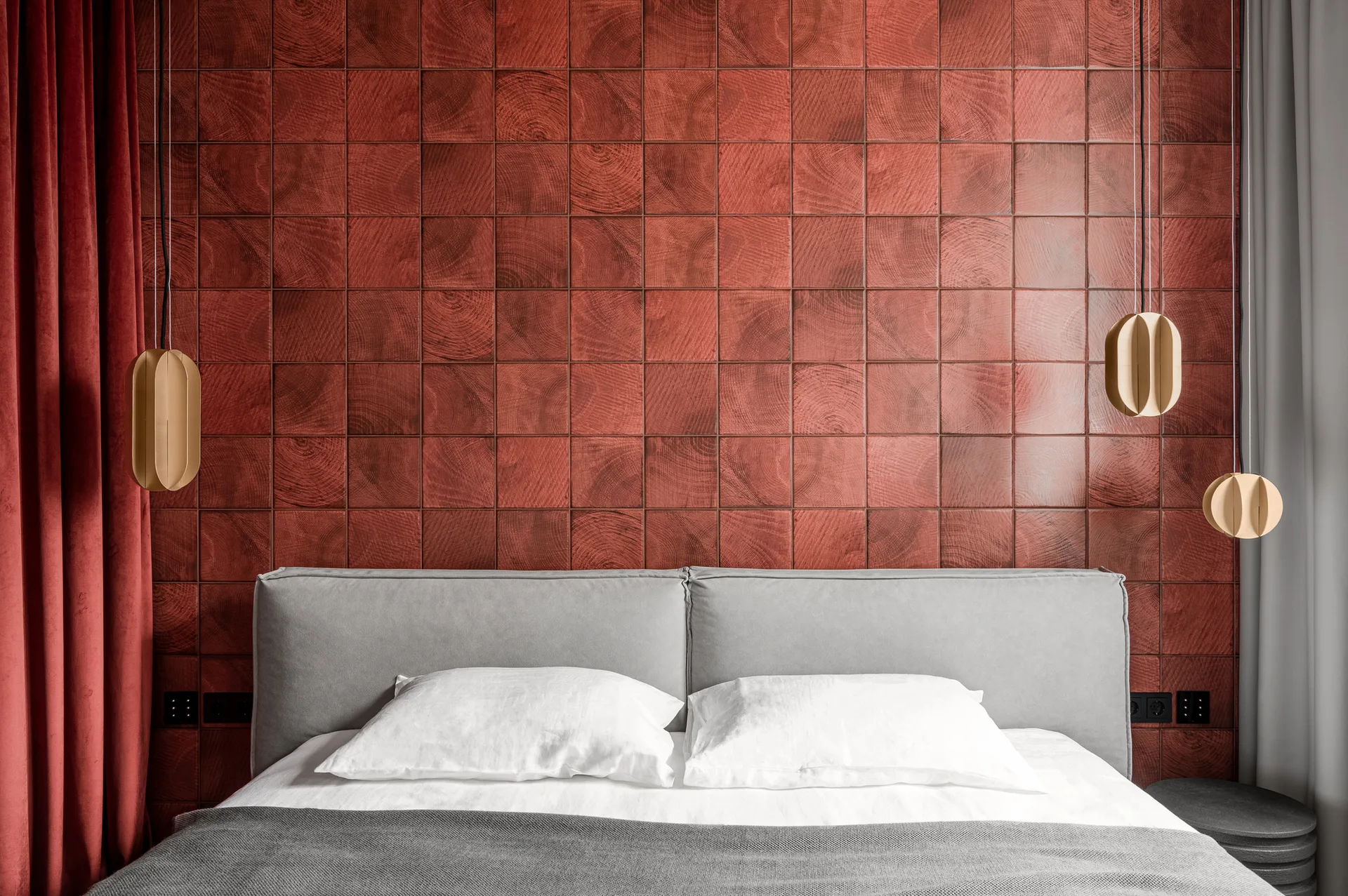 A grey upholstered bed is set against a wall of red square wood tiles with swirling grain patterns, flanked by modern gold pendant lights.