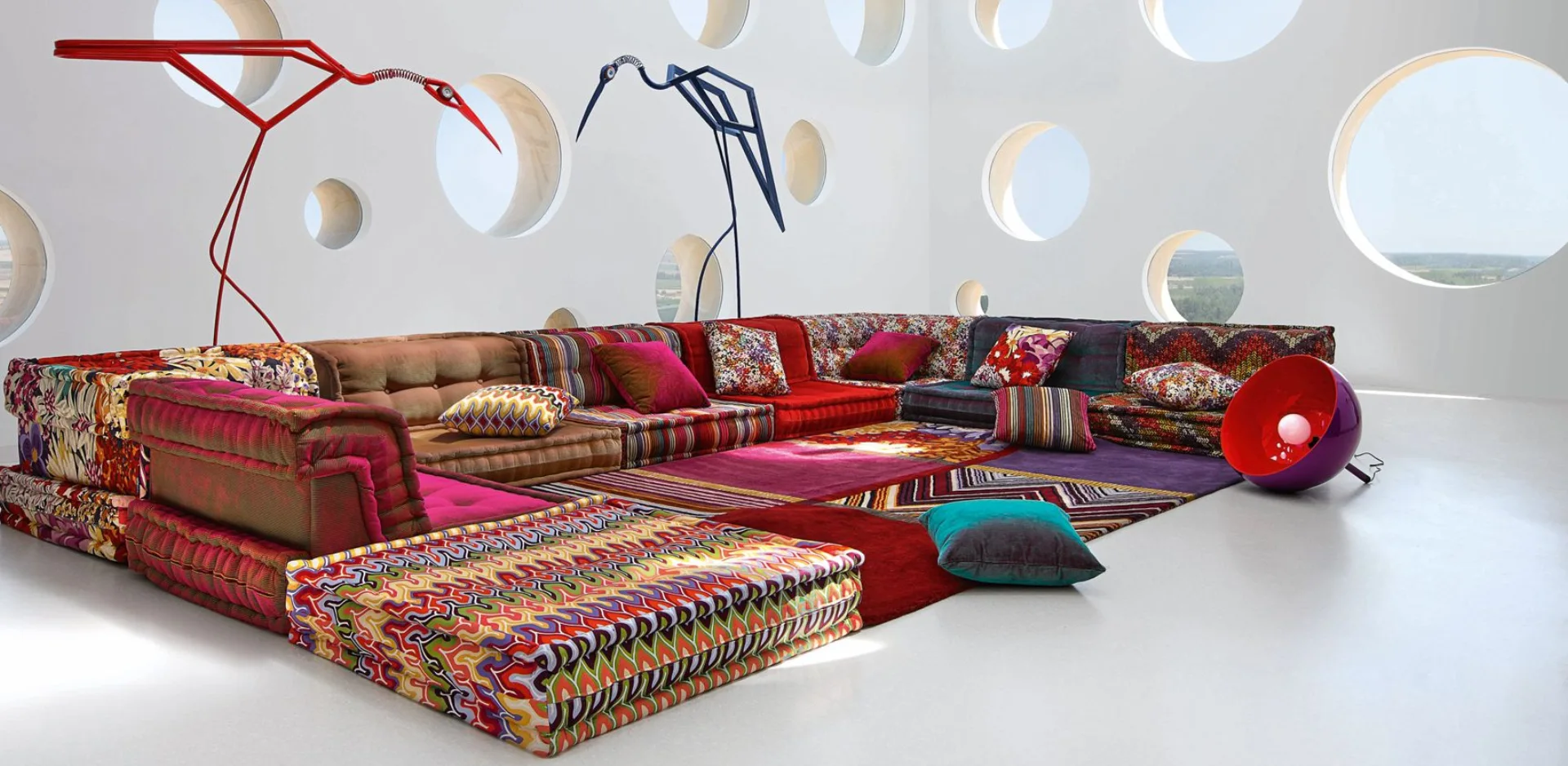 A spacious white room features a colorful, low-profile modular sectional sofa on a patterned rug next to a wall with multiple circular windows.