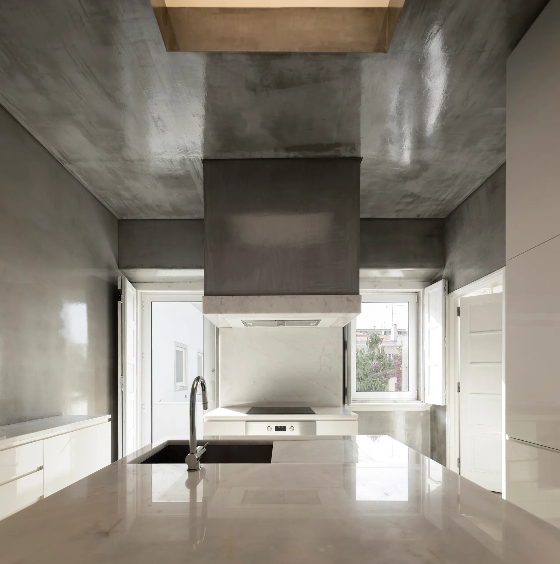 A modern kitchen featuring a polished stone island with a faucet, glossy grey plaster walls and ceiling, a large central range hood, and white cabinetry.