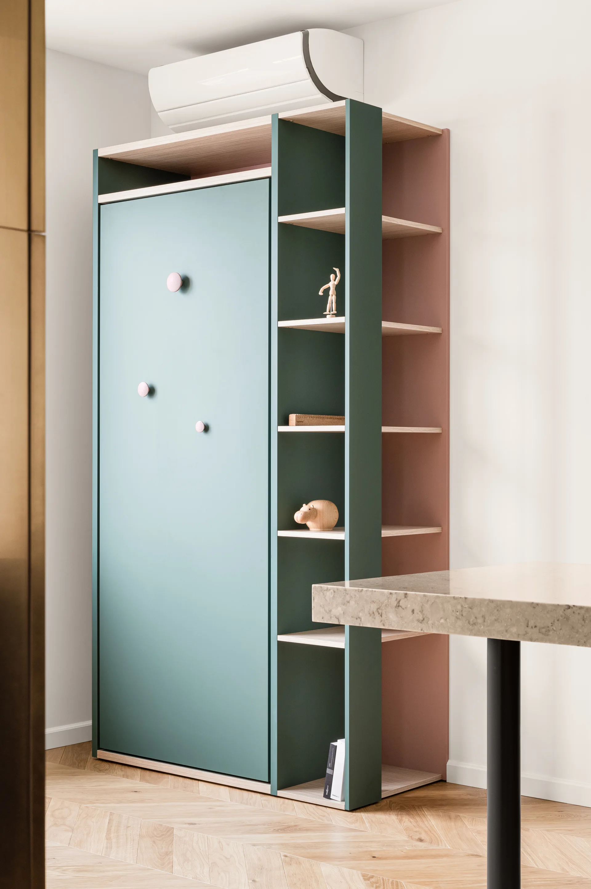 A tall modern cabinet featuring sage green panels and muted pink shelving stands on a herringbone pattern wood floor next to a stone-look table.
