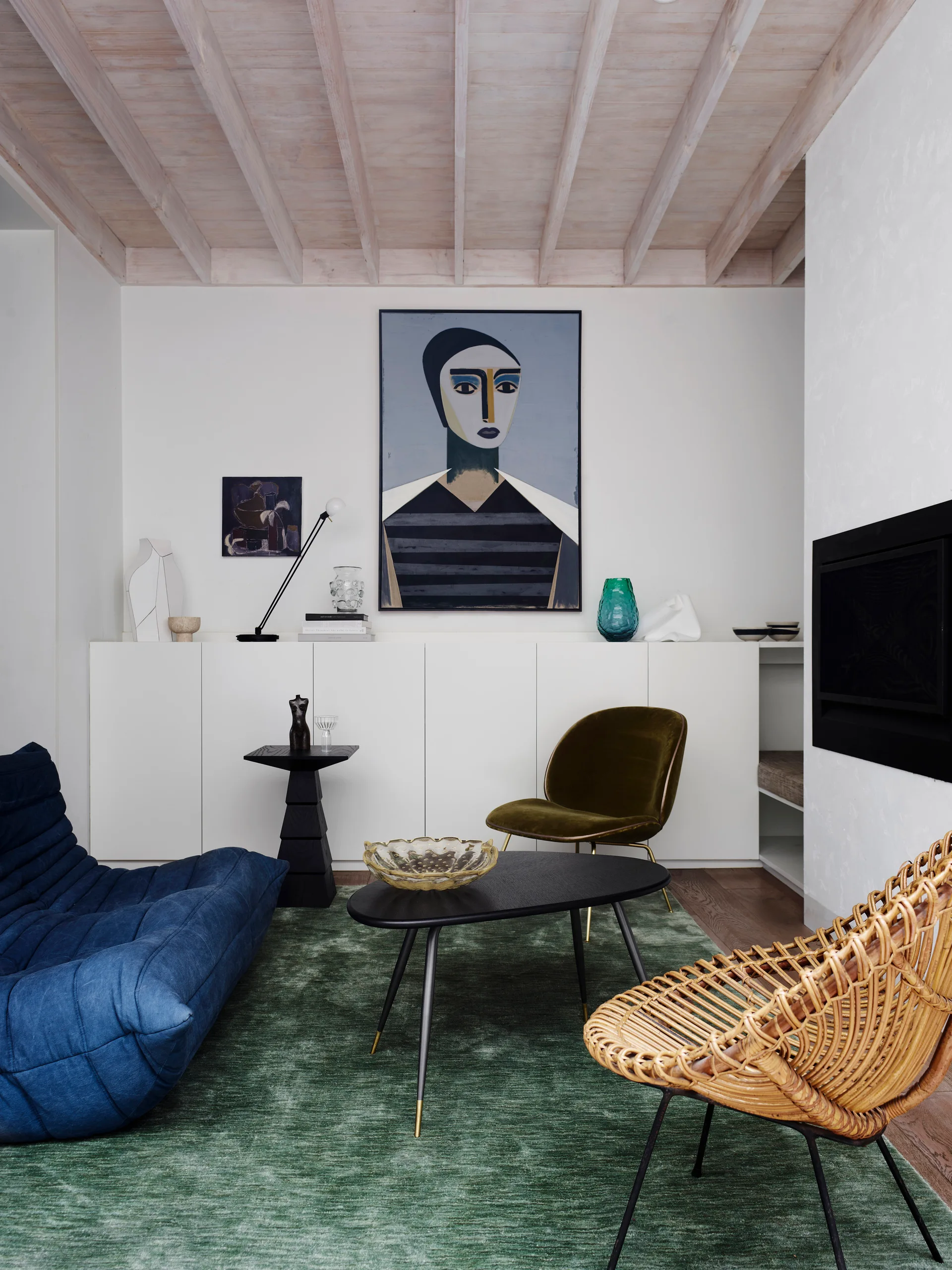 An eclectic living room featuring a blue tufted sofa, a woven rattan chair, a green area rug, and an exposed wood beam ceiling.