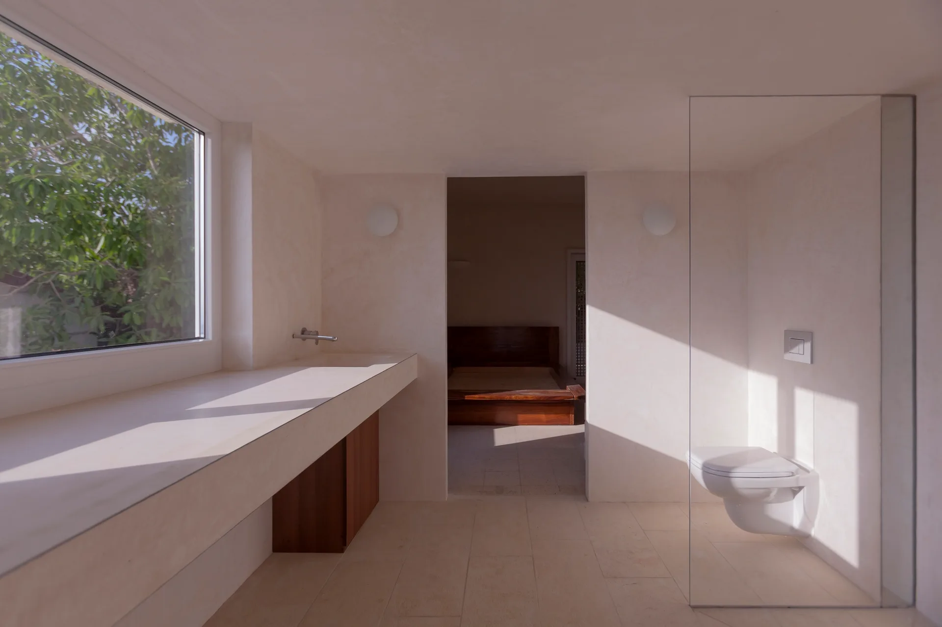 A minimalist sunlit bathroom with off-white plaster walls, a long integrated vanity counter, and a wall-mounted toilet behind a glass partition.
