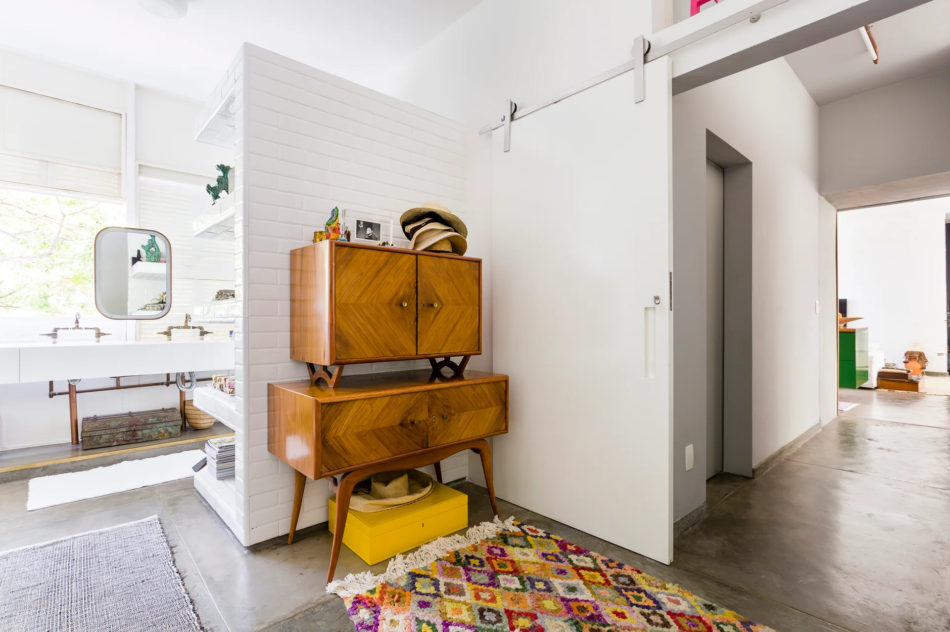 A stacked mid-century wooden cabinet sits next to a white sliding door on concrete floors with a colorful geometric rug.