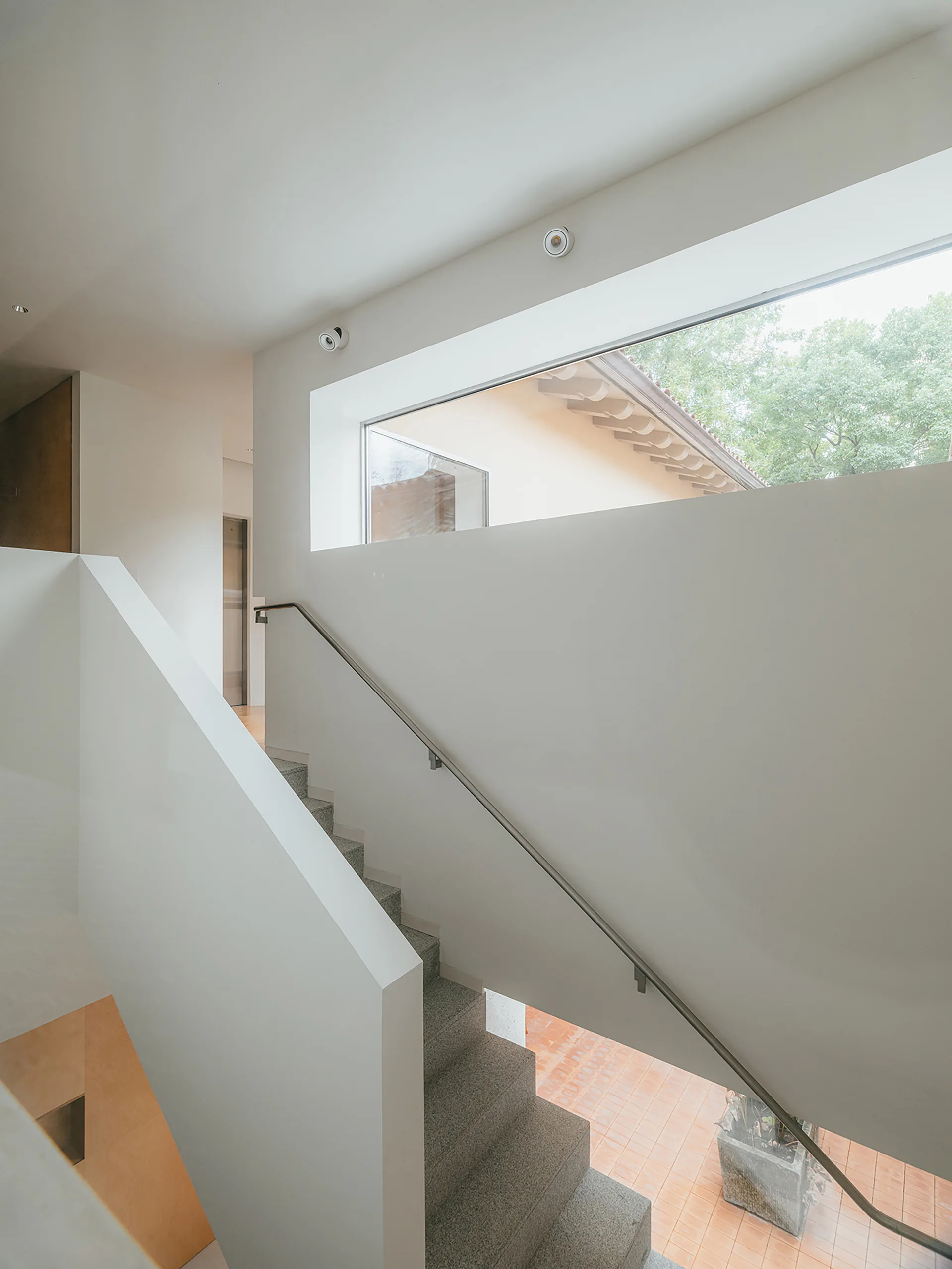 A minimalist white stairwell features textured stone stairs, a slim metal handrail, and a wide horizontal window framing outdoor trees.