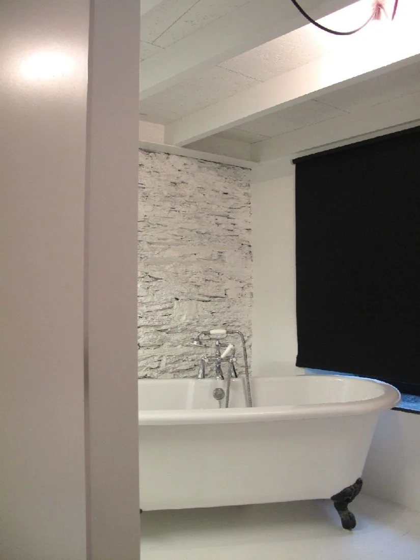A white claw-foot bathtub sits against a white painted stone wall below exposed ceiling beams next to a black window blind.