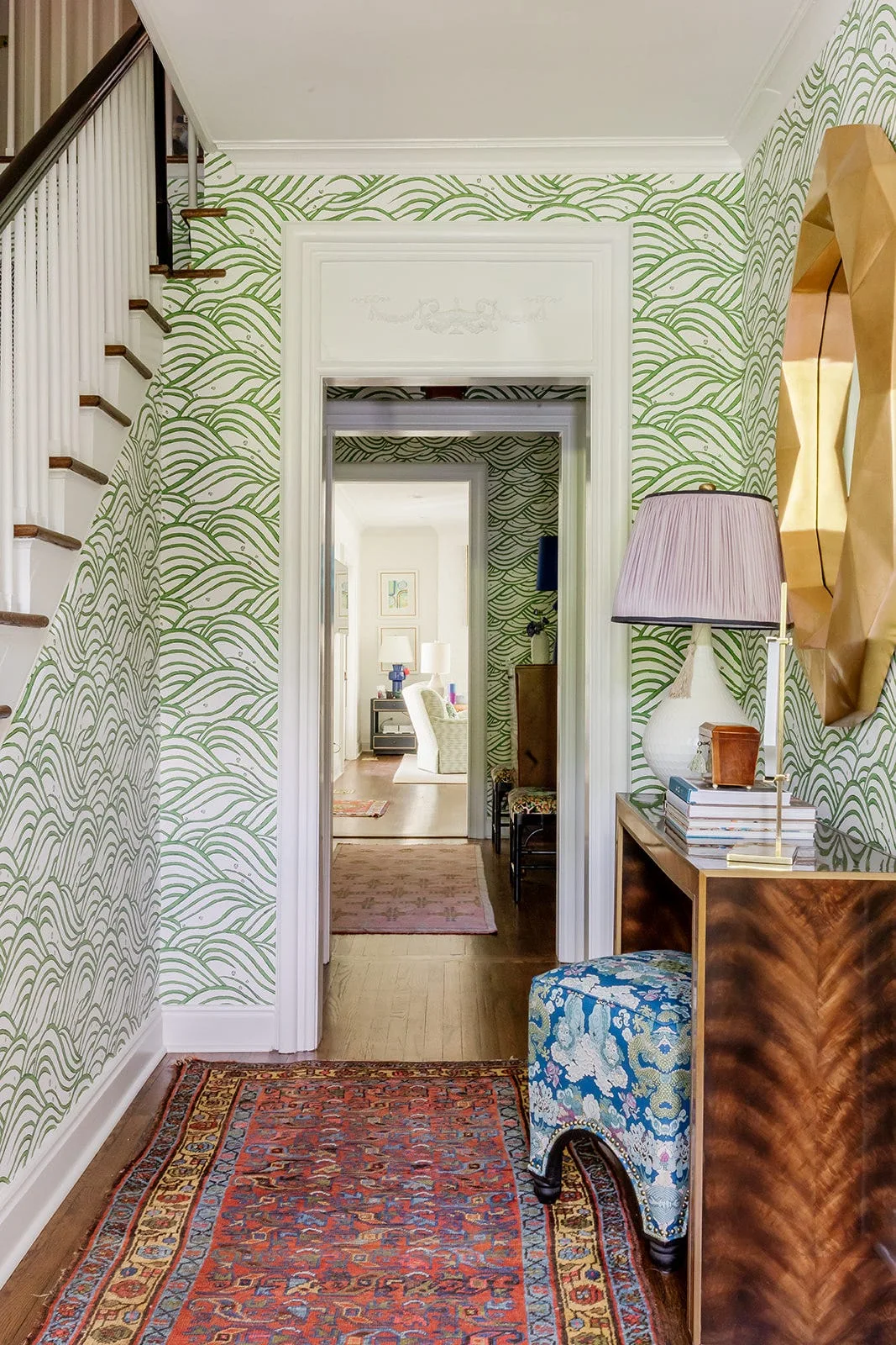 An entryway featuring green and white wave-patterned wallpaper, a wooden console table with a blue upholstered stool, and a patterned red area rug.