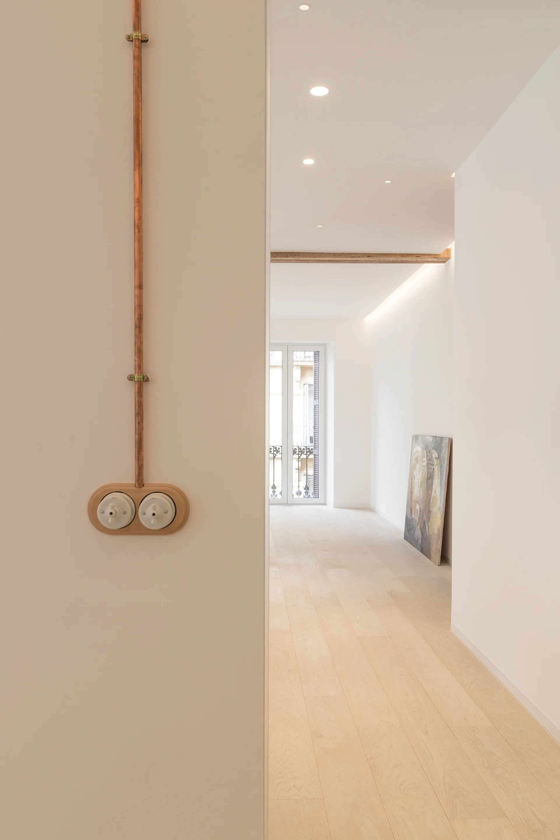 A minimalist white hallway with light wood flooring features exposed copper piping connected to a vintage light switch.