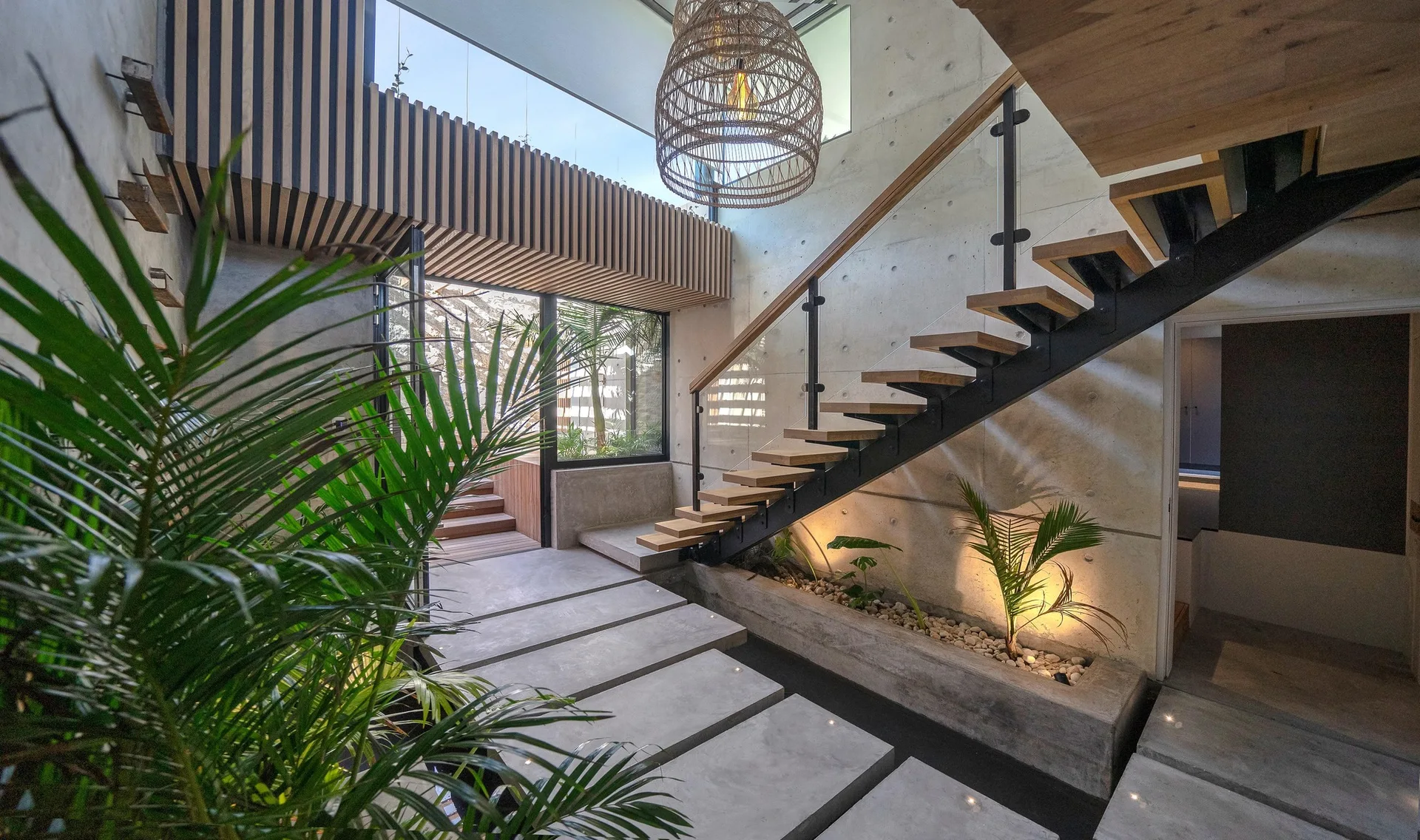 A modern concrete entryway features floating wooden stairs over an integrated concrete planter with plants and a large woven pendant light.