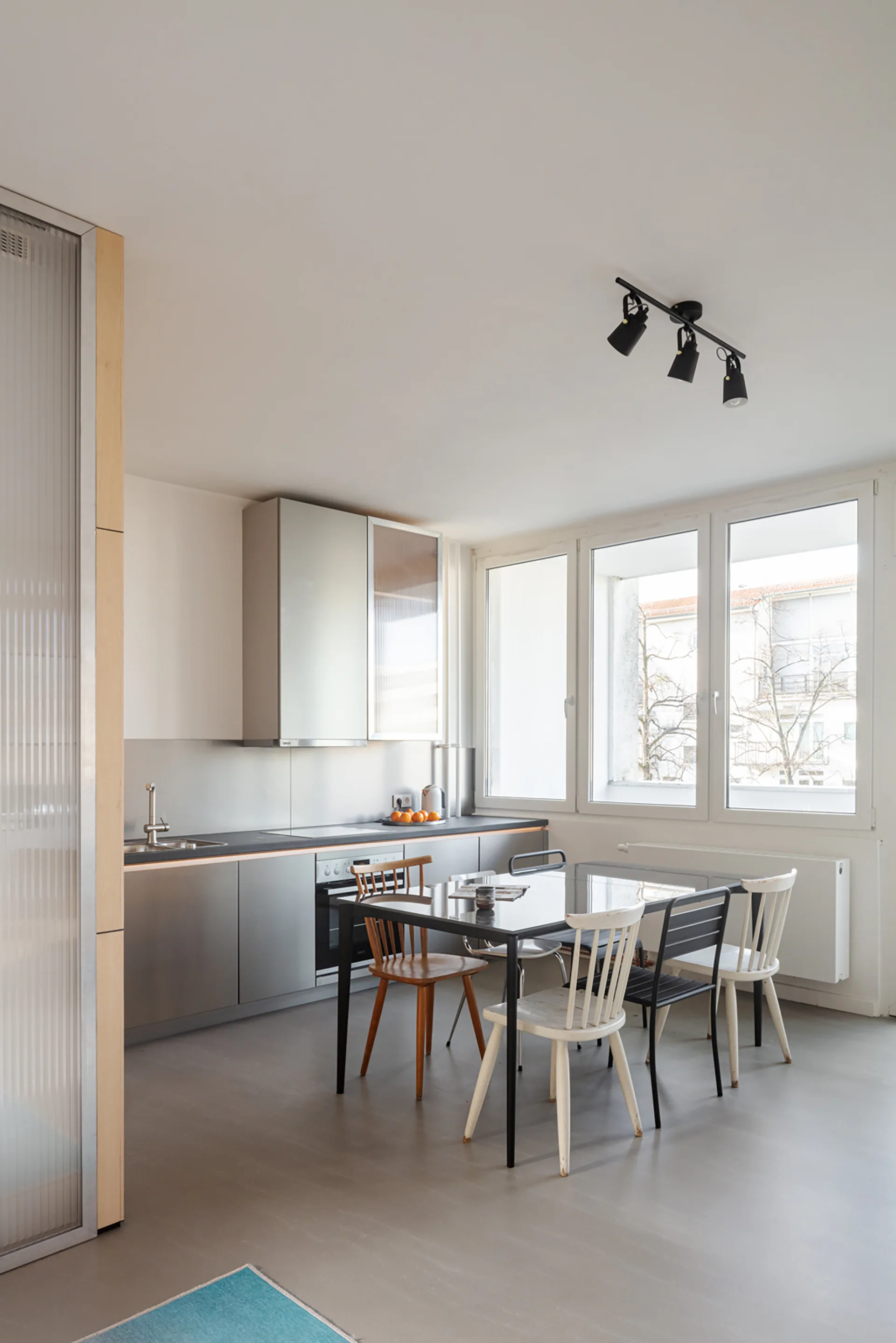 A contemporary kitchen and dining space featuring brushed metallic cabinets, a dark glass table, and an assortment of mismatched chairs.