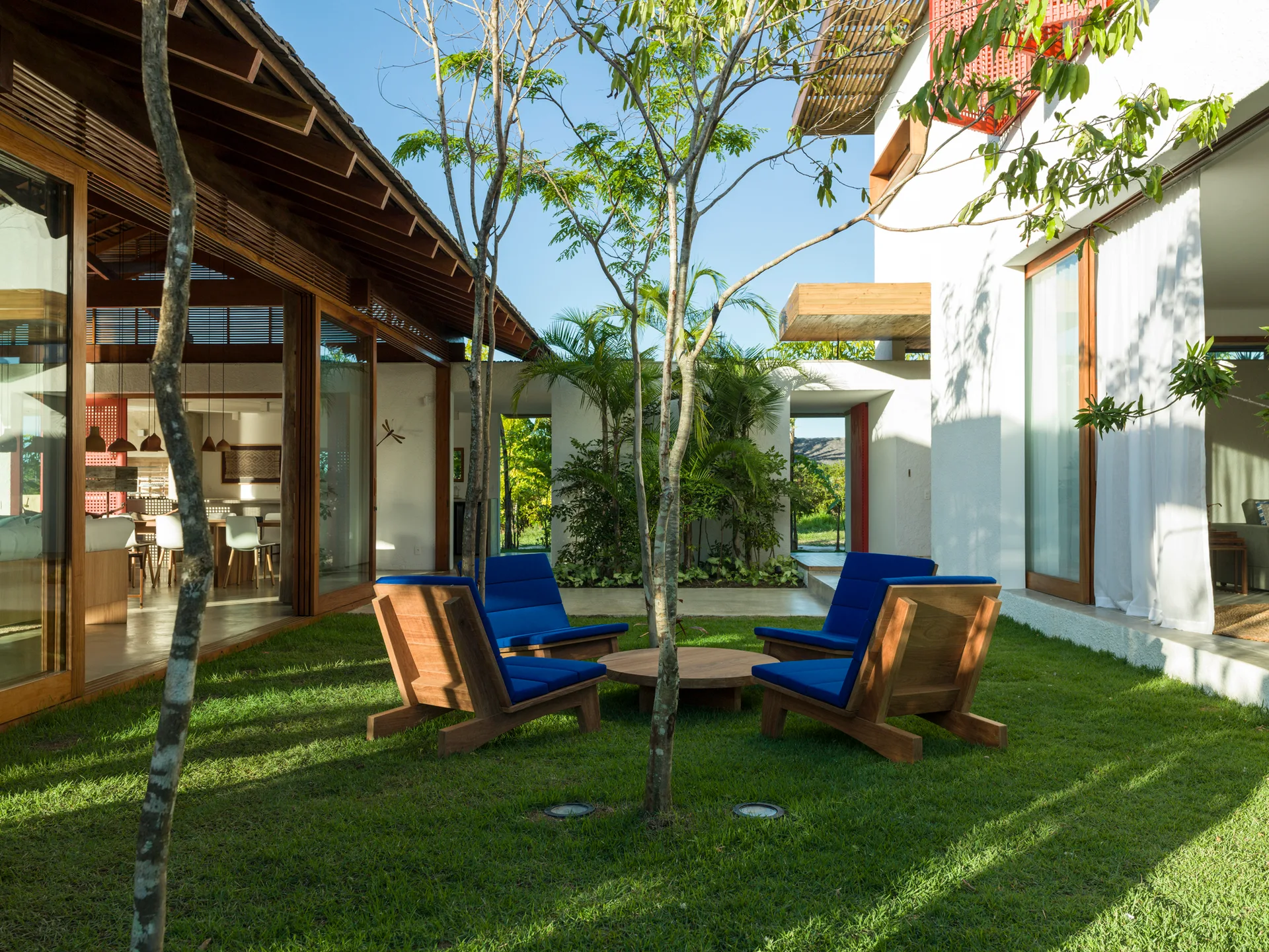 Blue-cushioned wooden lounge chairs are arranged around a low circular table on a grassy lawn in a modern courtyard surrounded by white walls.