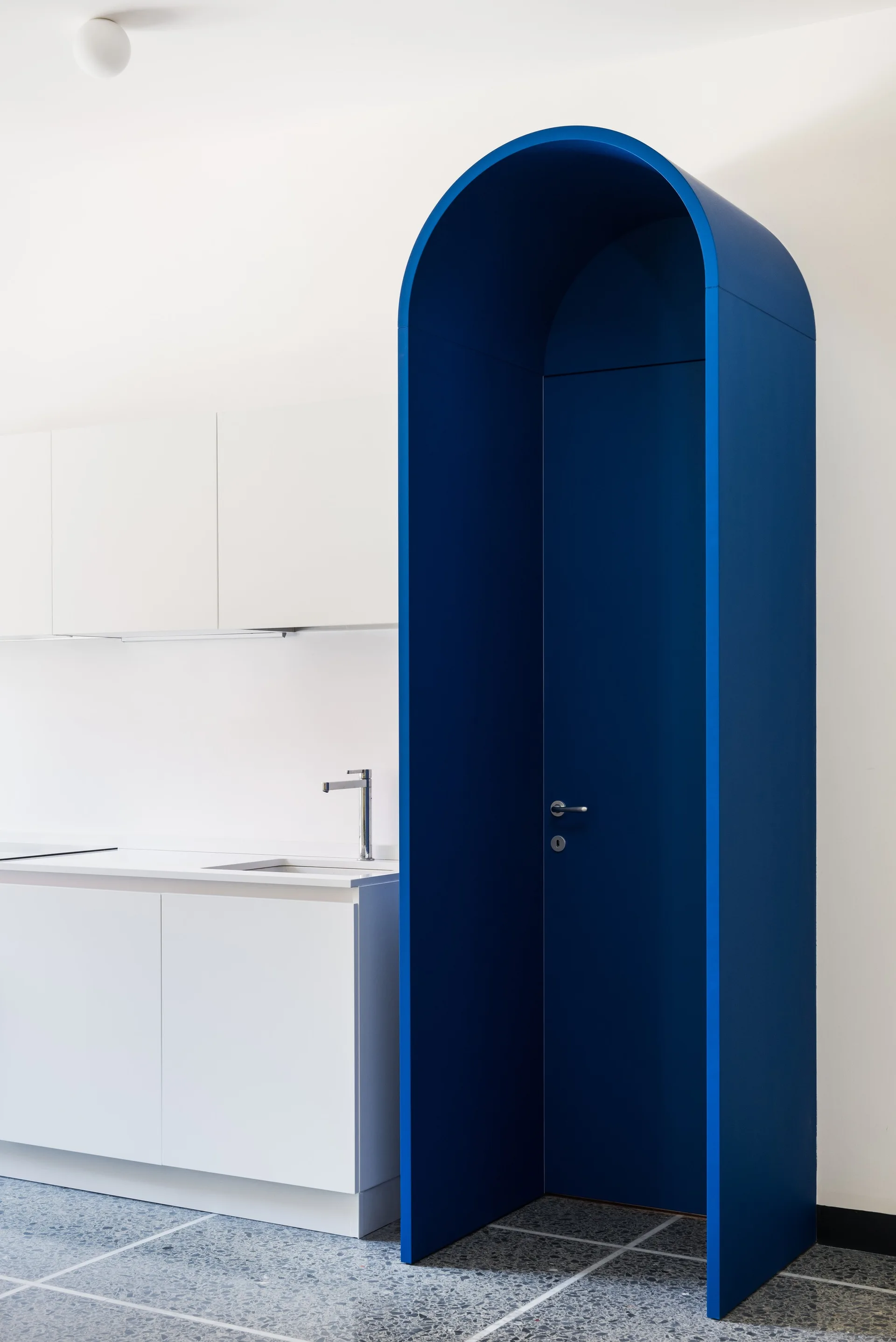 A minimalist white kitchen features flat cabinets, a modern faucet, terrazzo tile flooring, and a prominent blue arched doorway structure.