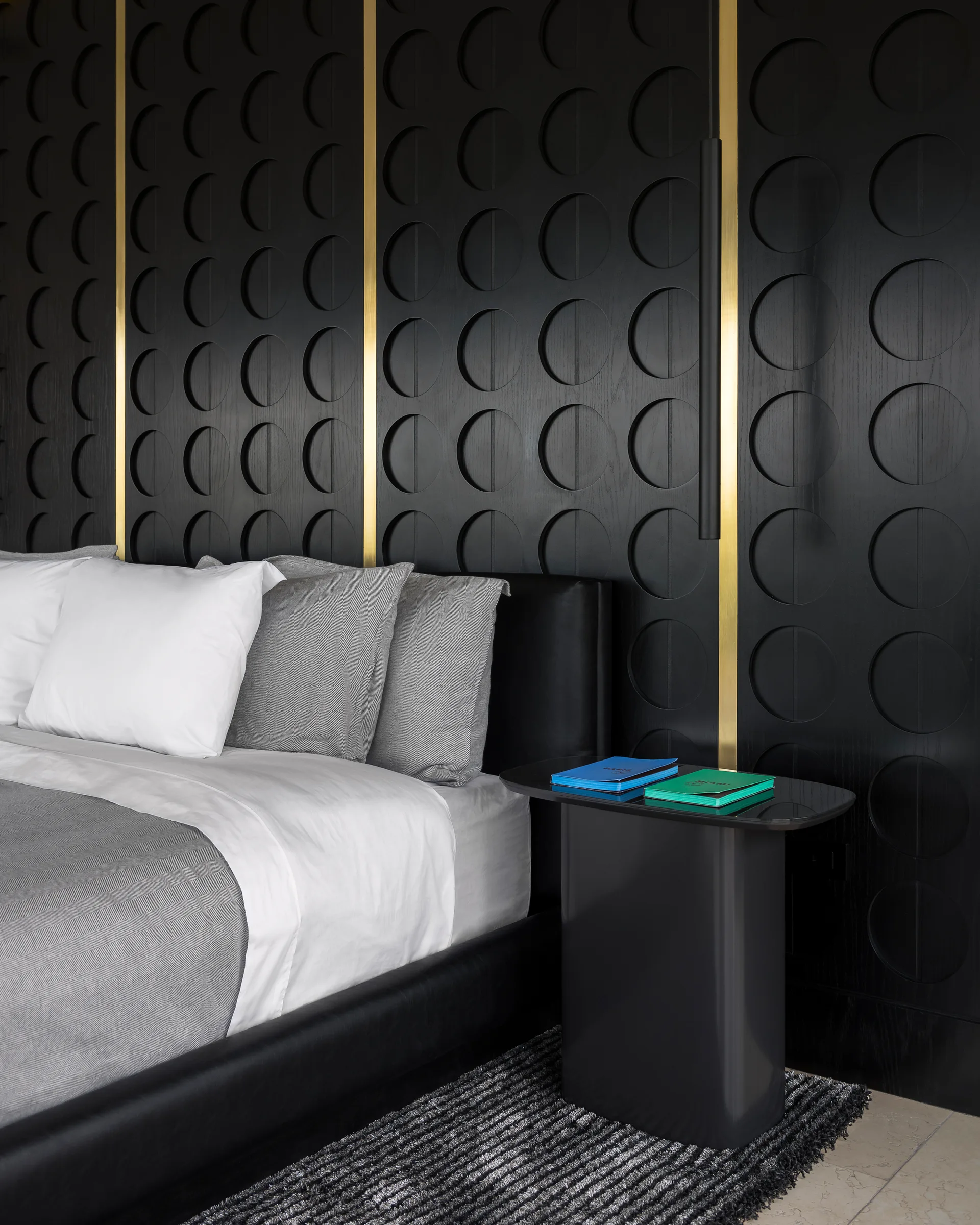 A bedroom featuring a black bed with grey bedding next to a black pedestal nightstand against a geometric patterned wall with vertical brass trim.