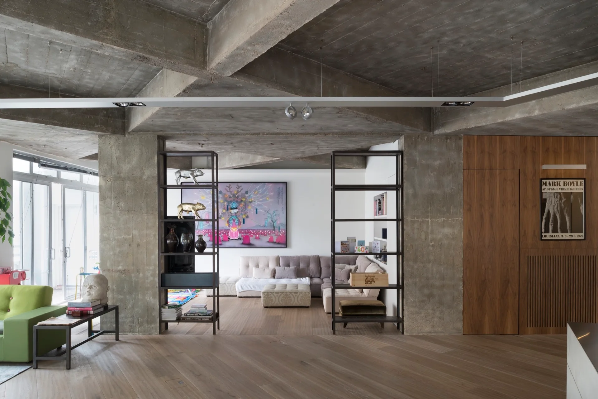An open-plan living room featuring exposed concrete ceilings and columns, light wood floors, and two metal bookshelves flanking a modular sofa.