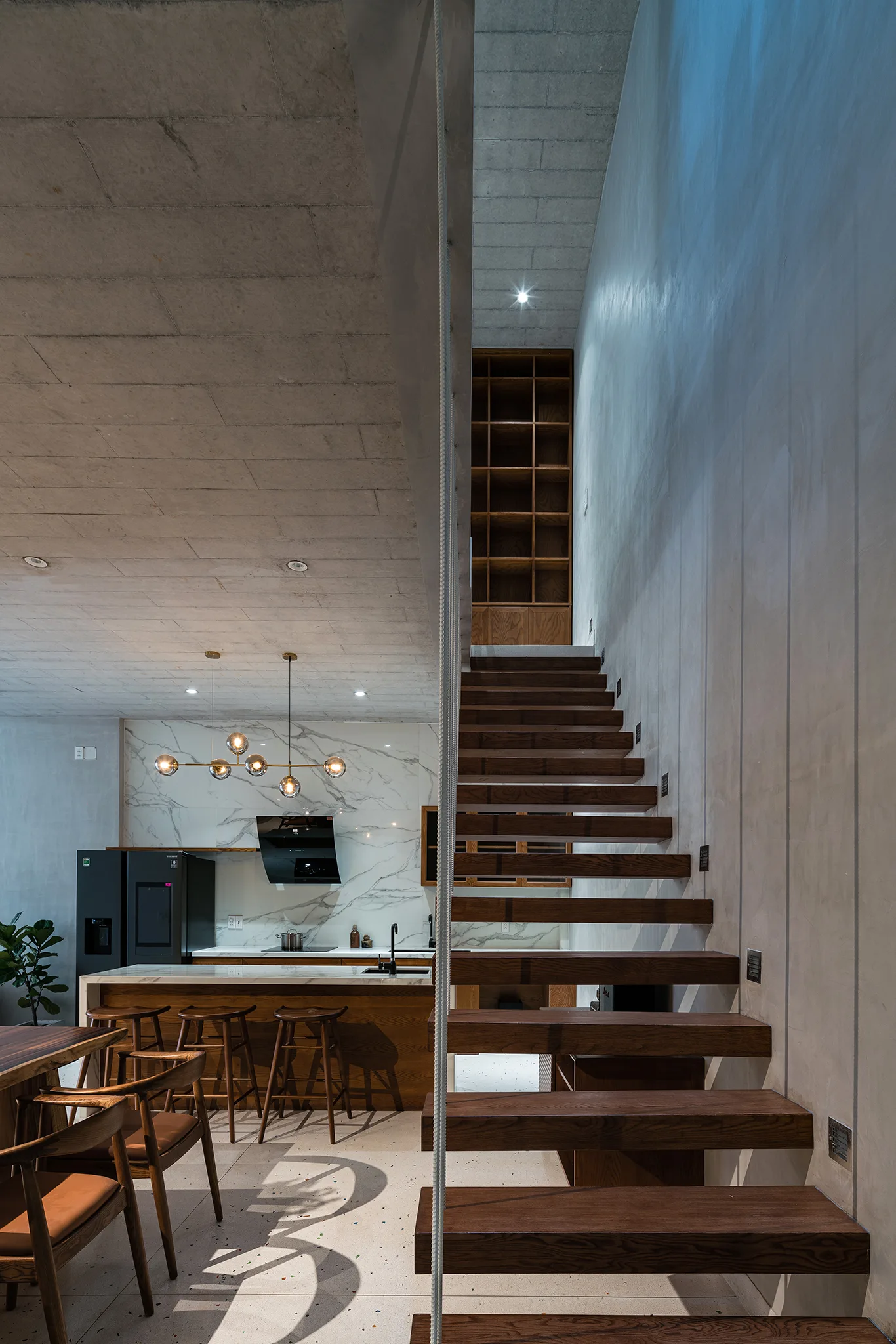 A floating wooden staircase rises beside a modern kitchen with a marble-topped island, wooden stools, and exposed concrete ceilings.