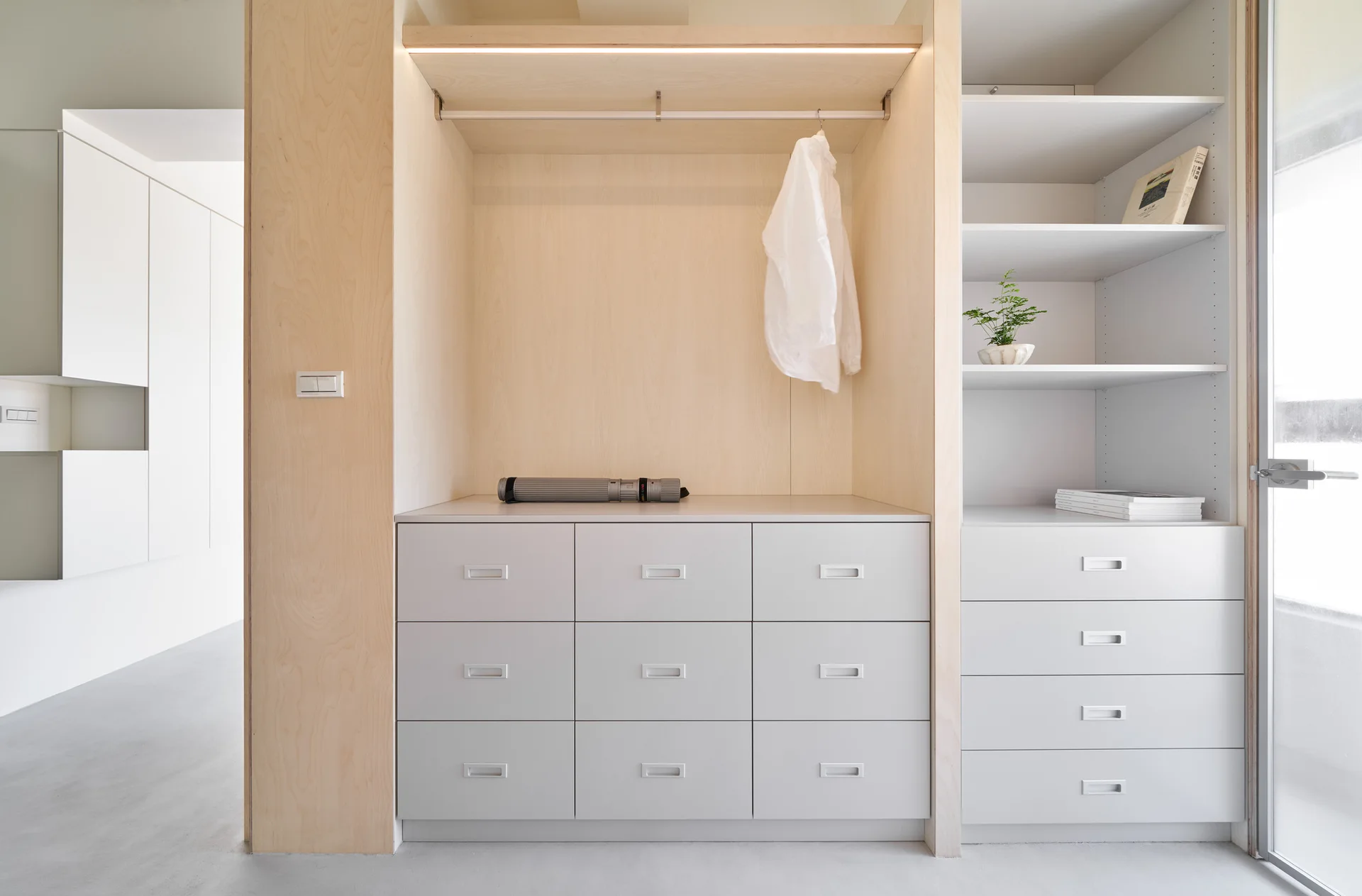 A minimalist built-in closet system features light wood framing, white drawer units with recessed pulls, and open shelving.
