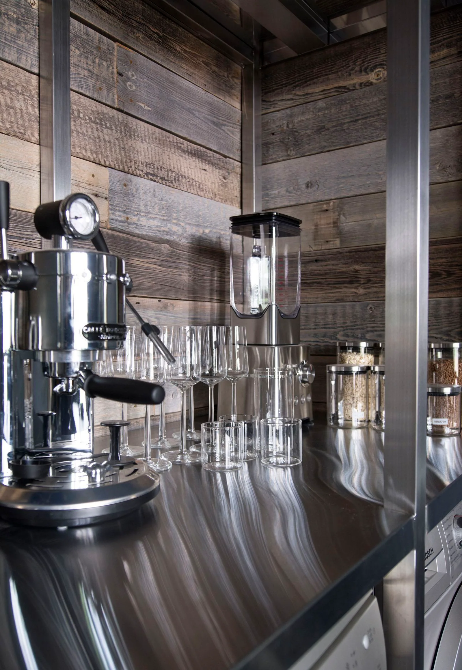 A chrome espresso machine, glassware, and glass jars sit on a stainless steel countertop against a rustic wall of weathered wood planks.