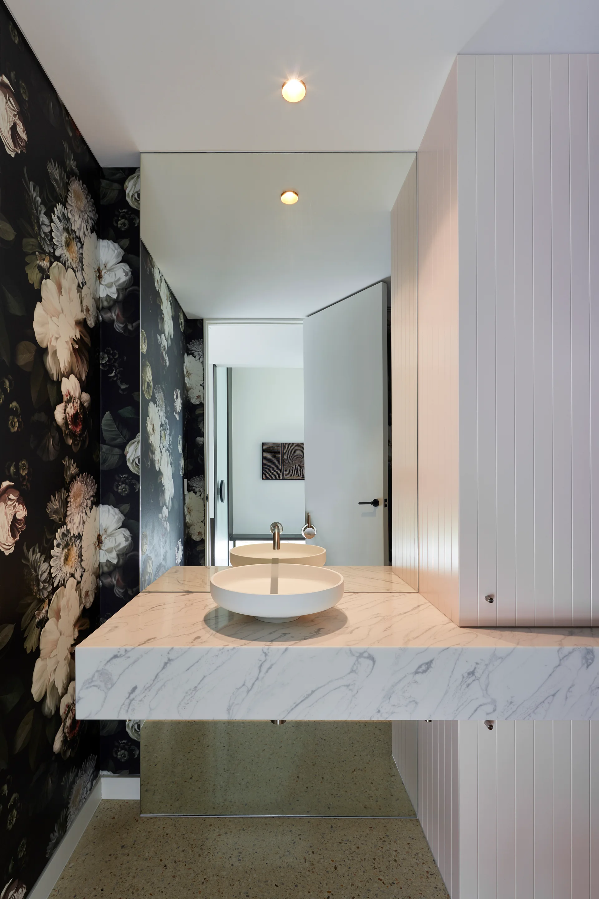 A modern powder room features a white marble floating vanity with a round vessel sink, dark floral wallpaper, and white vertical wood paneling.