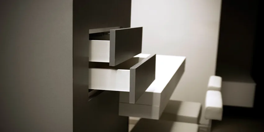 A close-up of a dark grey storage cabinet featuring two open drawers with white interiors, set against a background of white minimalist stools.