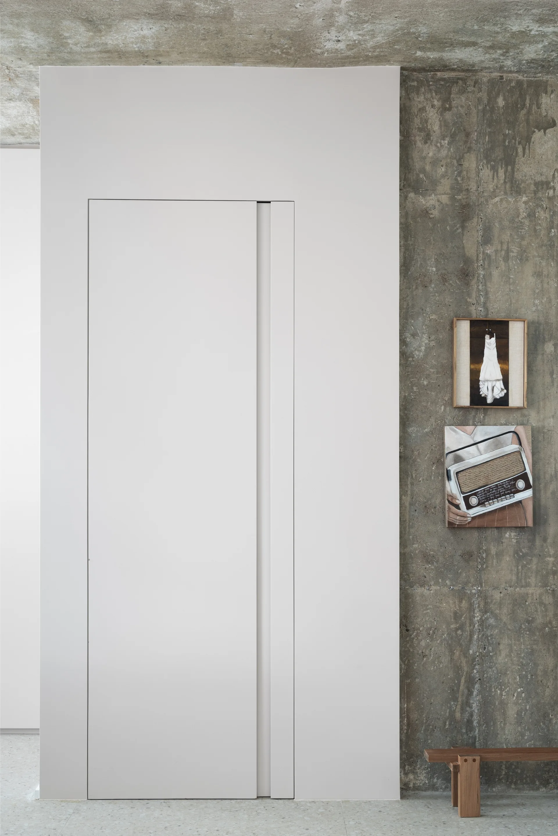 A minimalist white flush door is integrated into a smooth wall next to an exposed concrete wall with framed art.