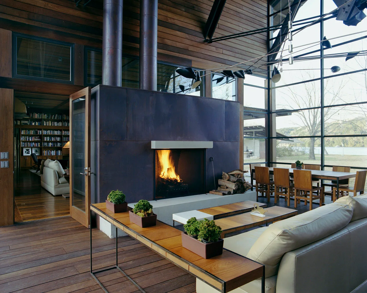A spacious cabin living room featuring a large dark metal fireplace, wood-paneled walls, and a floor-to-ceiling glass wall looking out at the water.