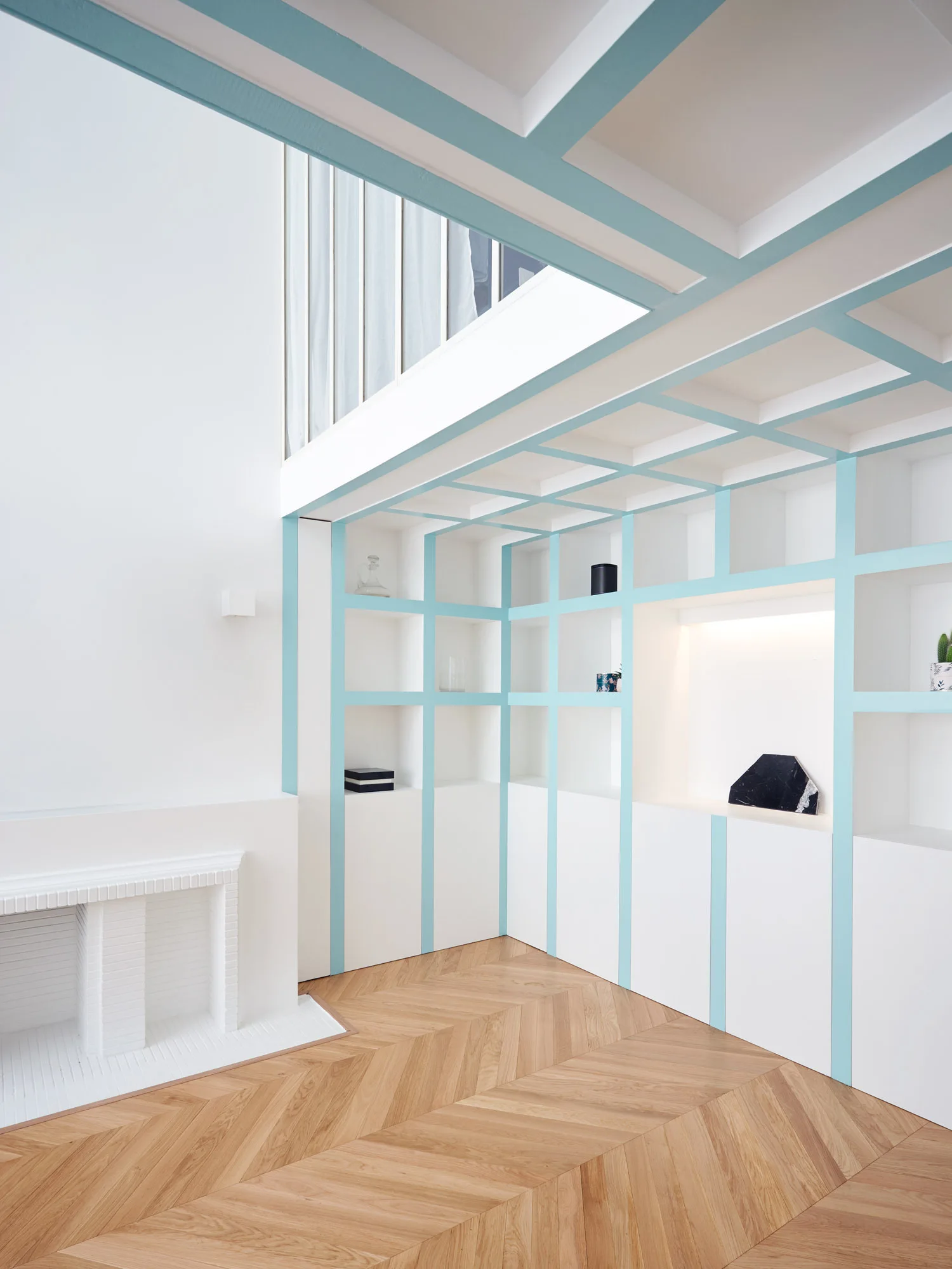 A bright interior with white grid built-in shelving painted with light blue trim, matching ceiling beams, and a natural wood herringbone floor.