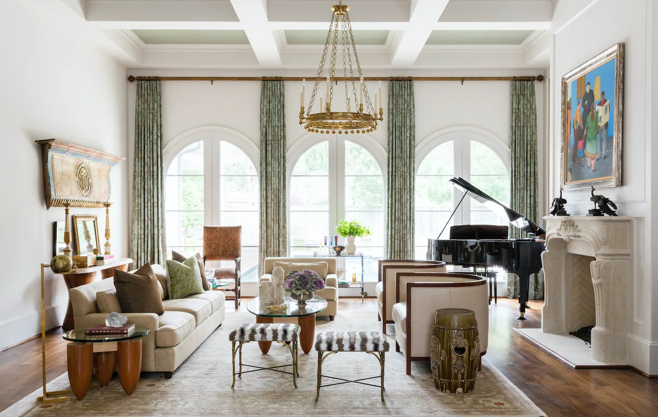 A formal living room with a grand piano, a stone fireplace, and light seating arranged before three arched windows with patterned drapery.
