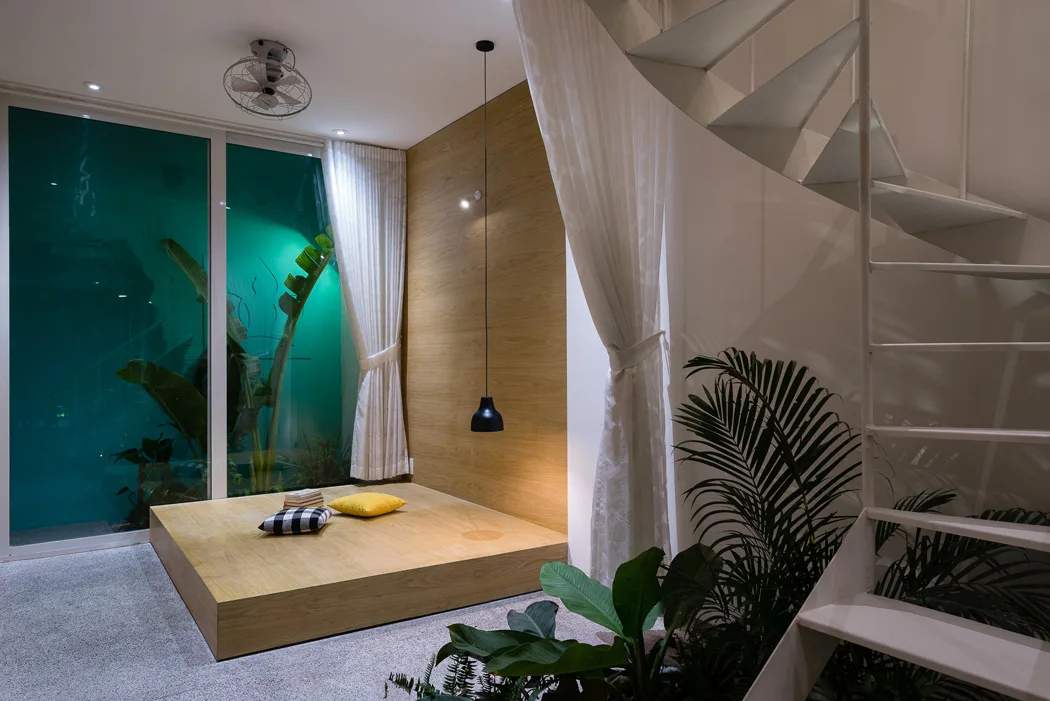 A minimalist interior space features an elevated wooden platform with cushions next to a white staircase and a large glass window.