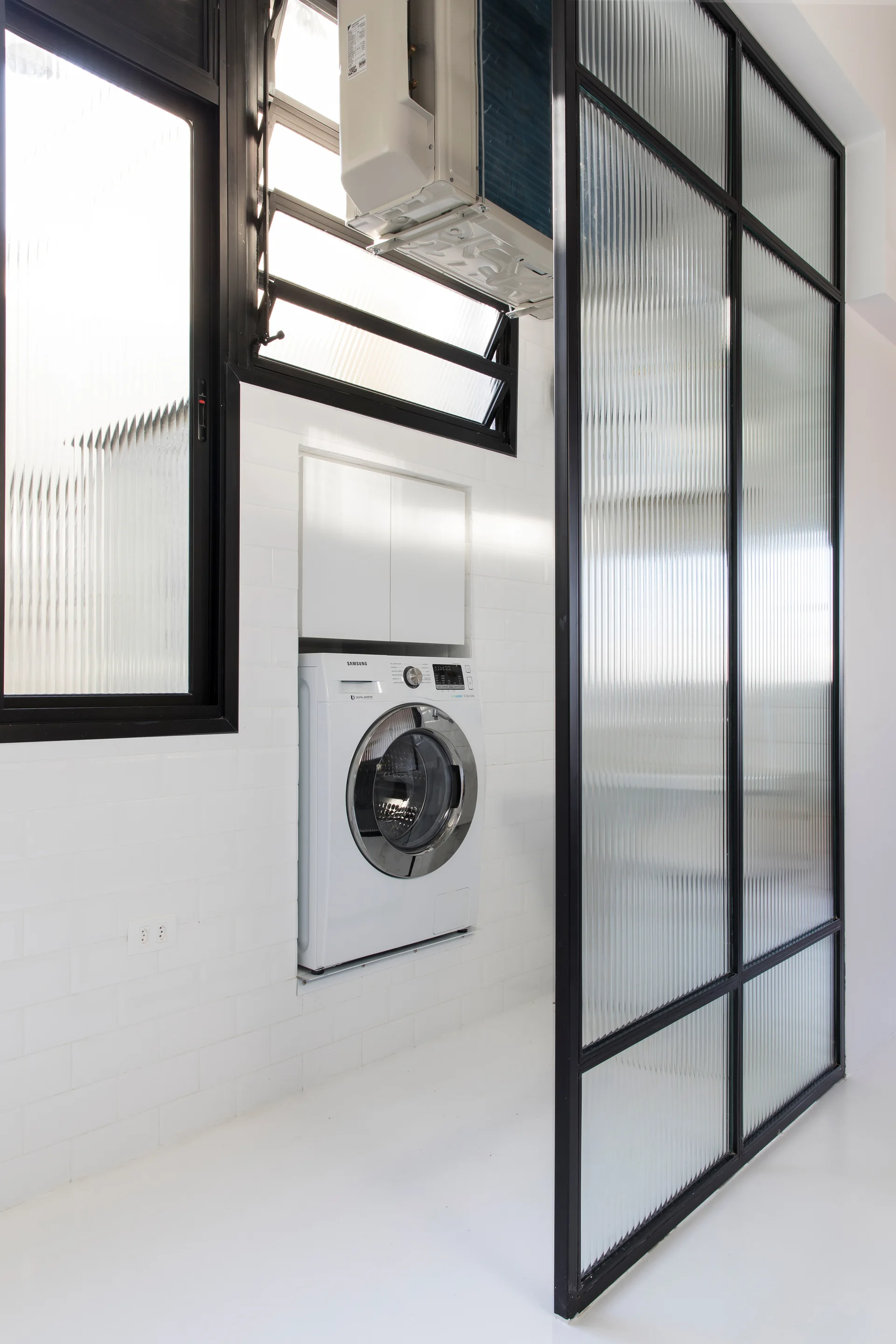 A white washing machine is built into a subway-tiled wall next to a large black-framed fluted glass partition in a modern utility room.