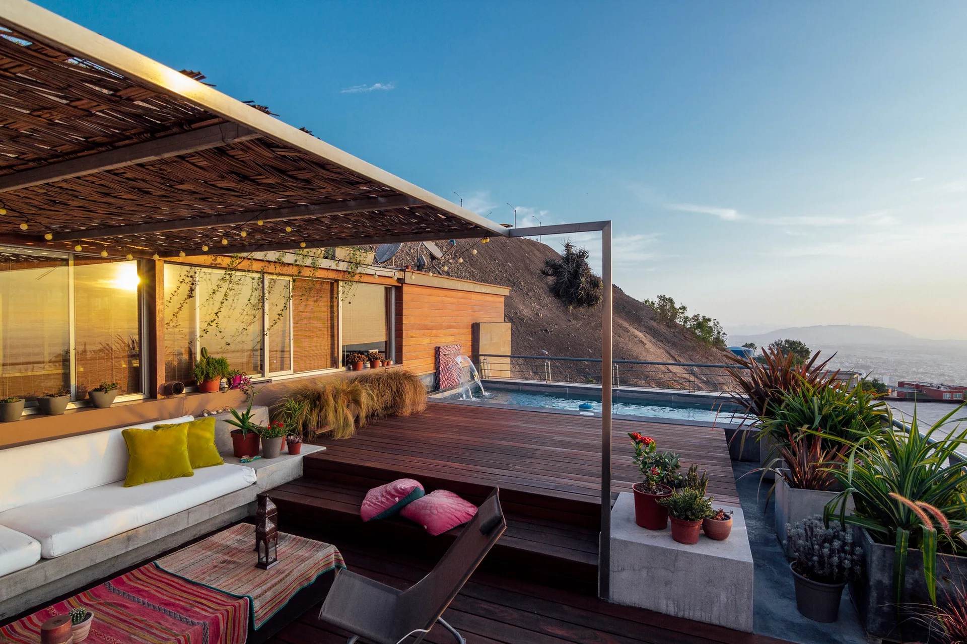 An outdoor hillside terrace features a multi-level wooden deck, a built-in concrete sofa under a thatched pergola, potted plants, and a small pool.