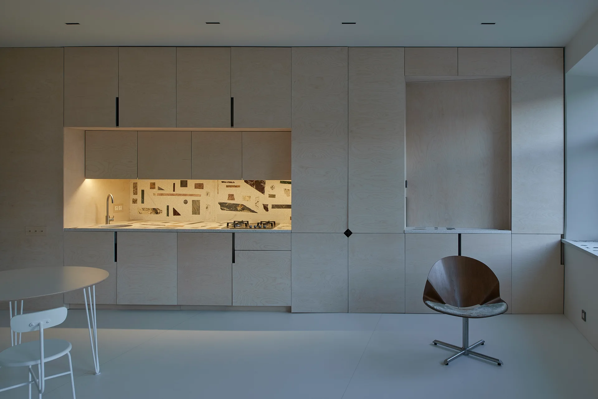 A modern kitchen featuring a full wall of light wood plywood cabinets, a sink niche with a geometric stone backsplash, and a dining chair.