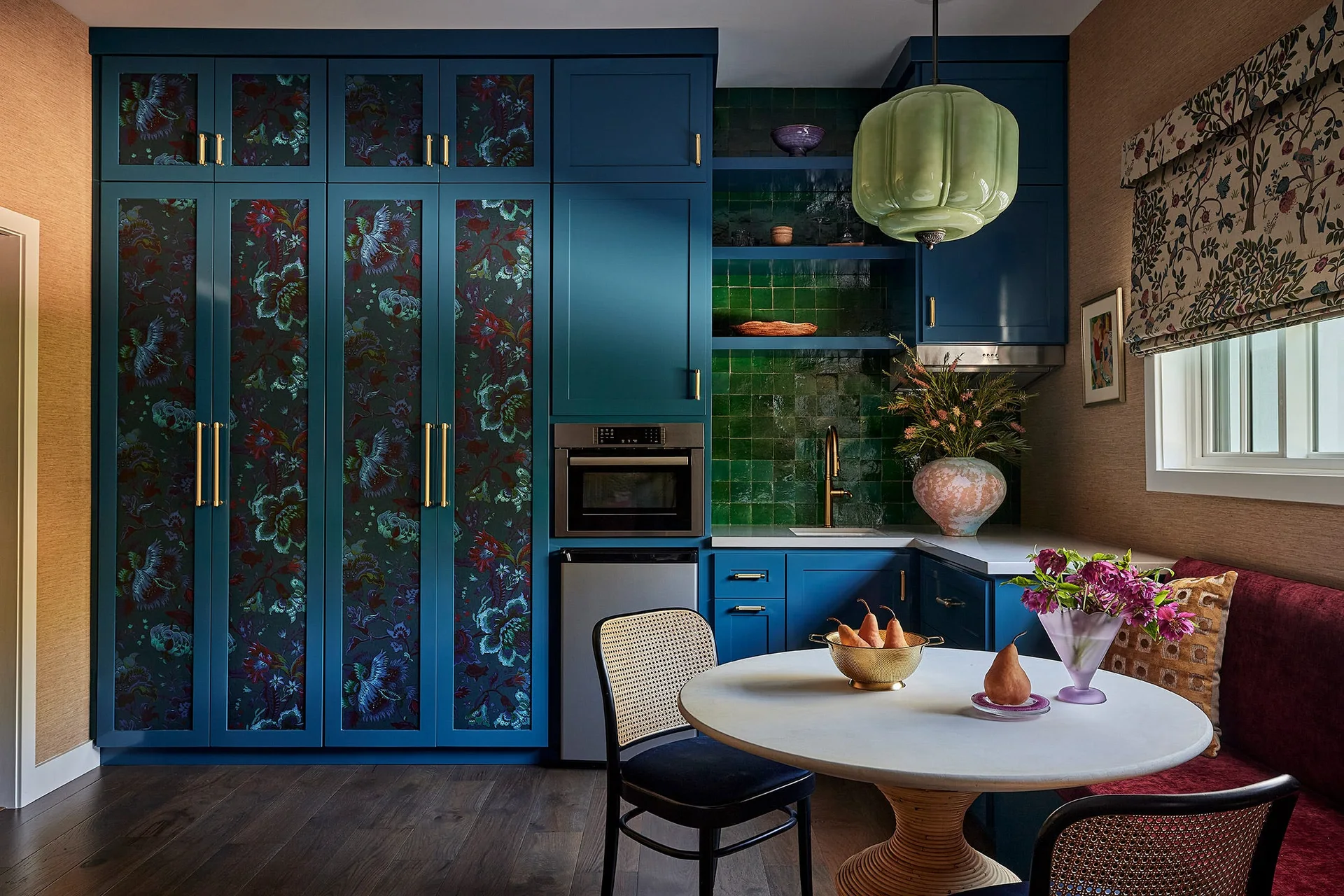 A kitchen dining nook featuring deep blue cabinetry with floral patterned door inserts, a green tiled backsplash, and a round pedestal table.