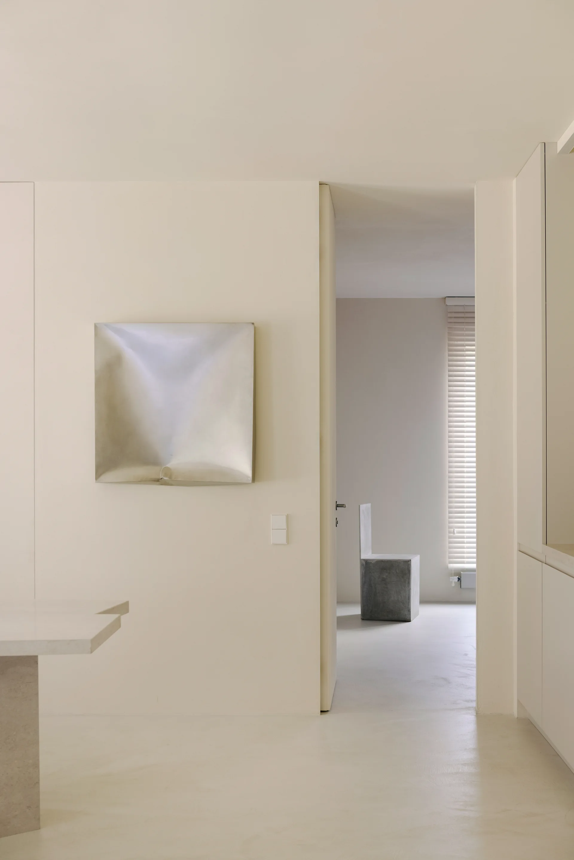 A minimalist beige hallway features a metallic square artwork on the wall and an open doorway leading to a room with a concrete chair.