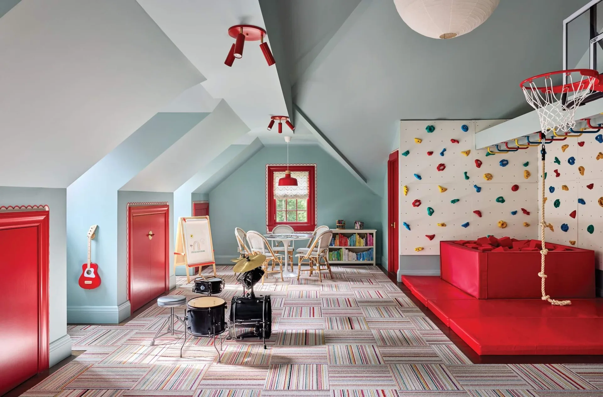 An attic playroom features light blue walls, a climbing wall with a red foam pit, a striped carpet, and built-in red doors.
