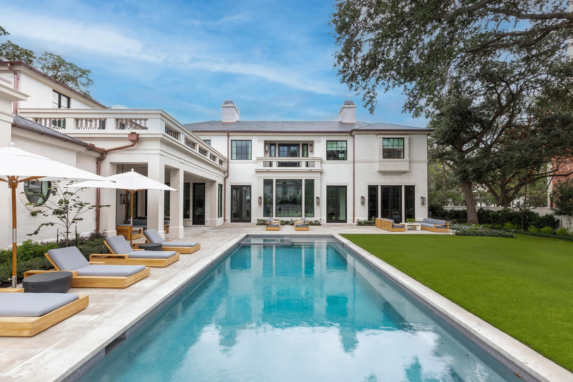 A large rectangular swimming pool is flanked by a stone patio with grey lounge chairs and a manicured green lawn behind a white stucco house.