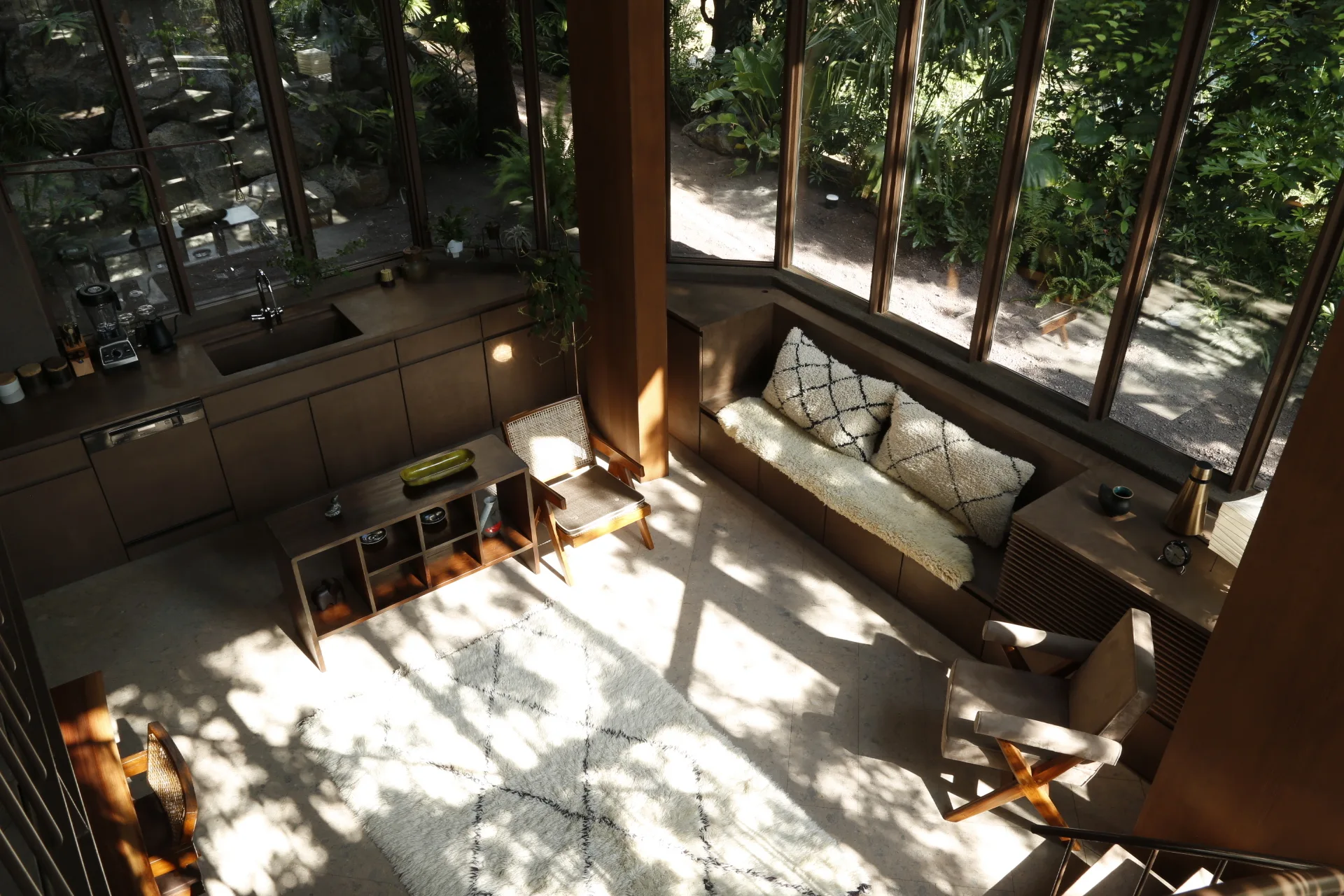 A sunlit living and kitchen area features dark wood cabinetry, a built-in bench under large glass windows, and textured cream rugs and pillows.