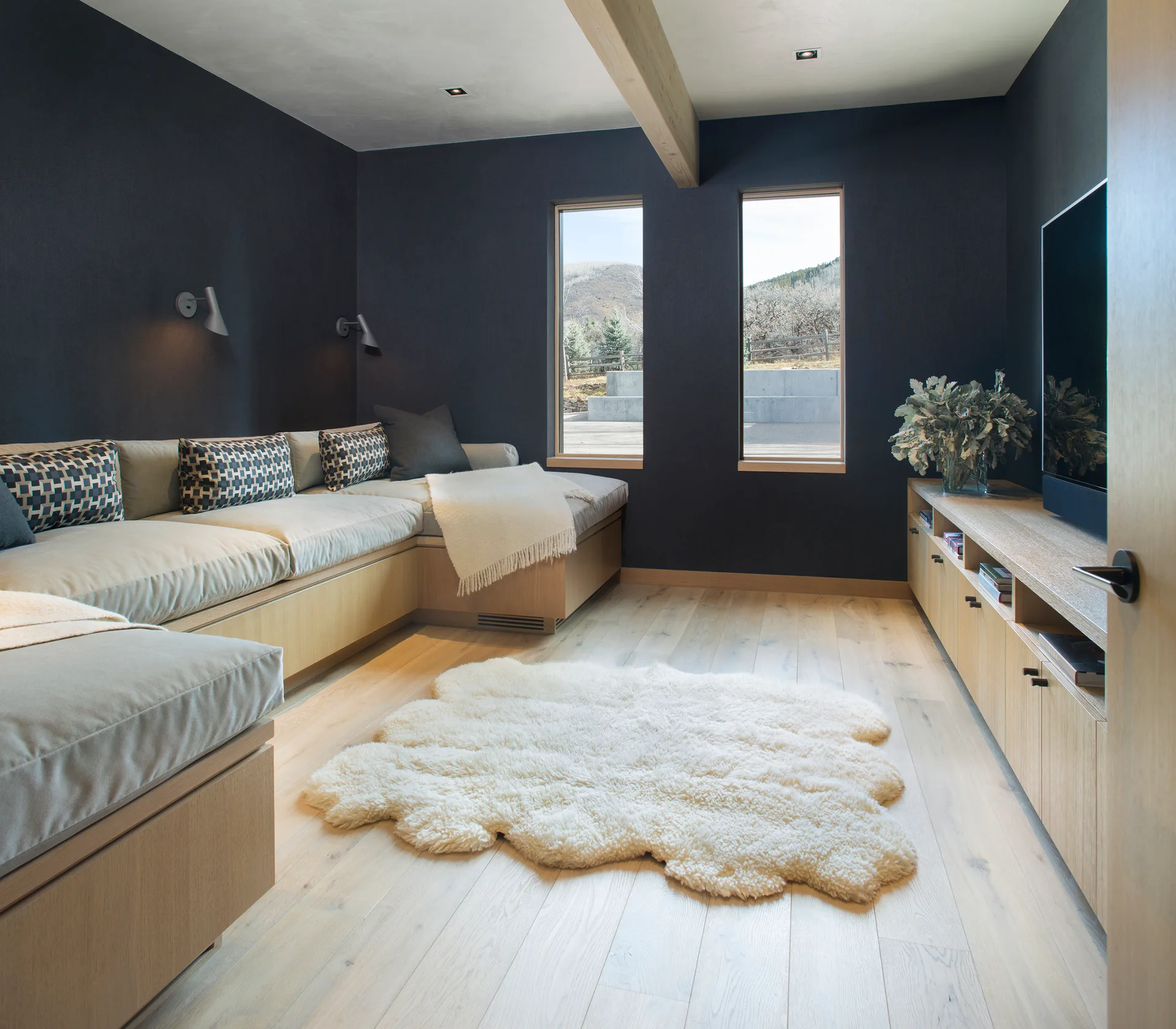 A cozy room features built-in light wood bench seating, dark charcoal walls, two narrow windows, a white sheepskin rug, and a matching wood console.