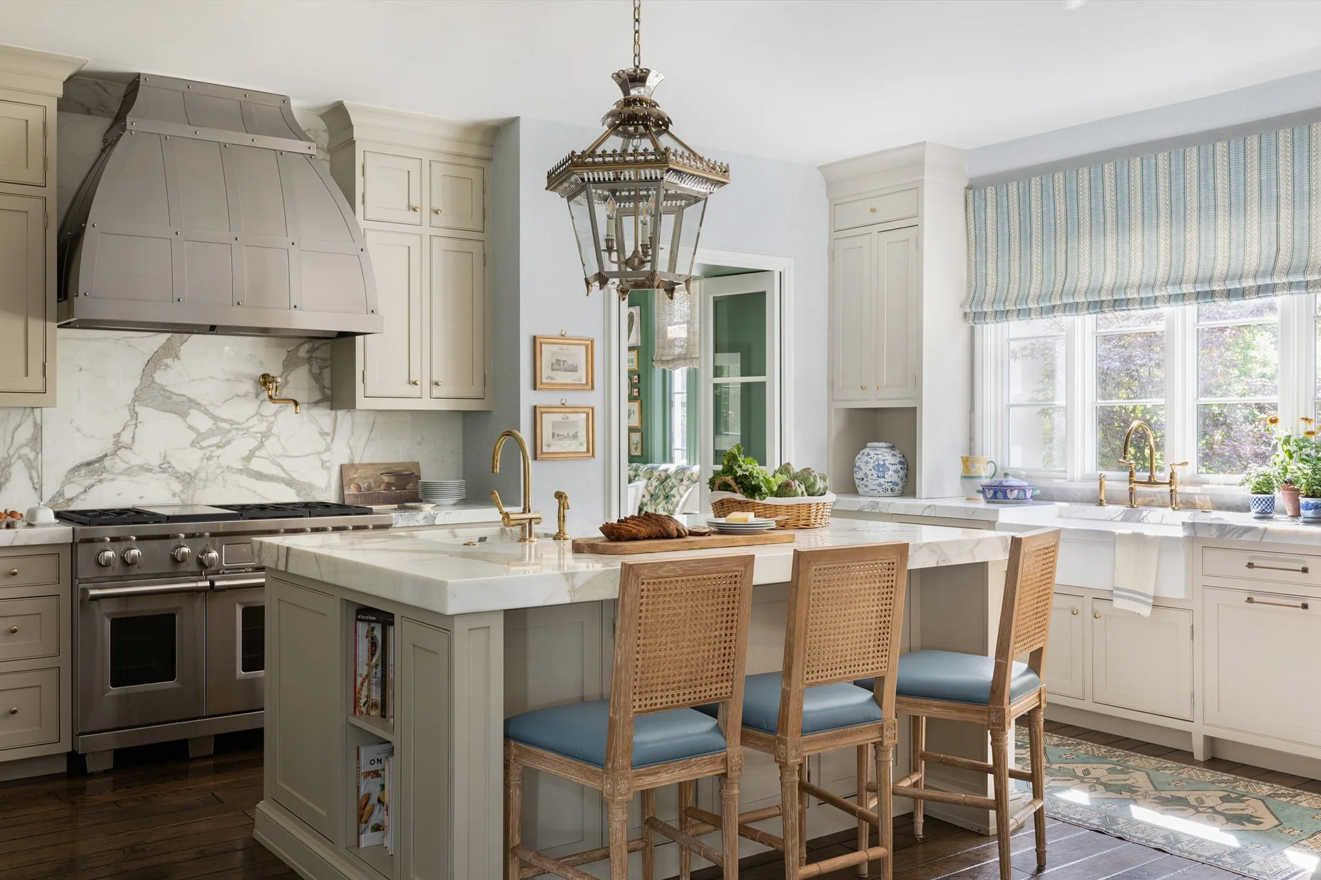 A traditional kitchen featuring a marble-topped island with three cane-back stools, cream cabinetry, a large metal range hood, and a lantern pendant.