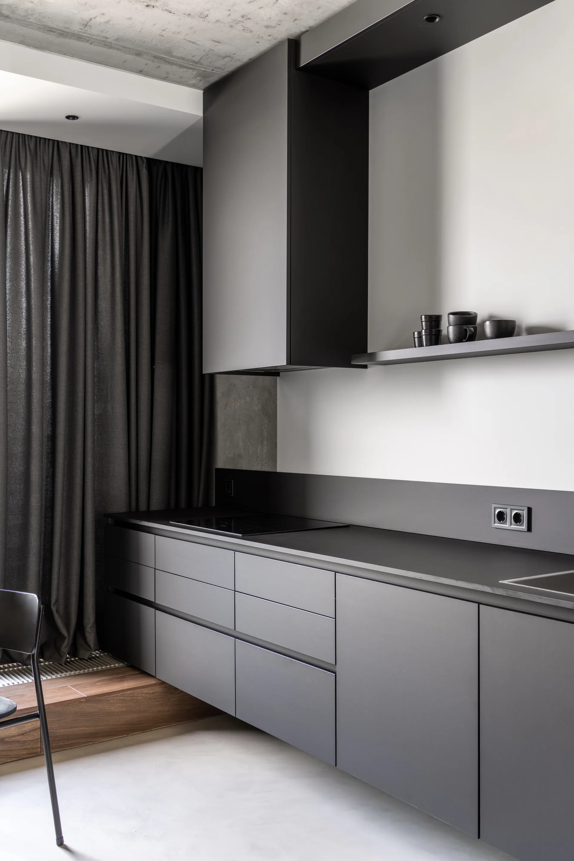 A minimalist kitchen features matte charcoal grey cabinetry, an integrated cooktop, a raw concrete ceiling, and dark floor-to-ceiling drapes.