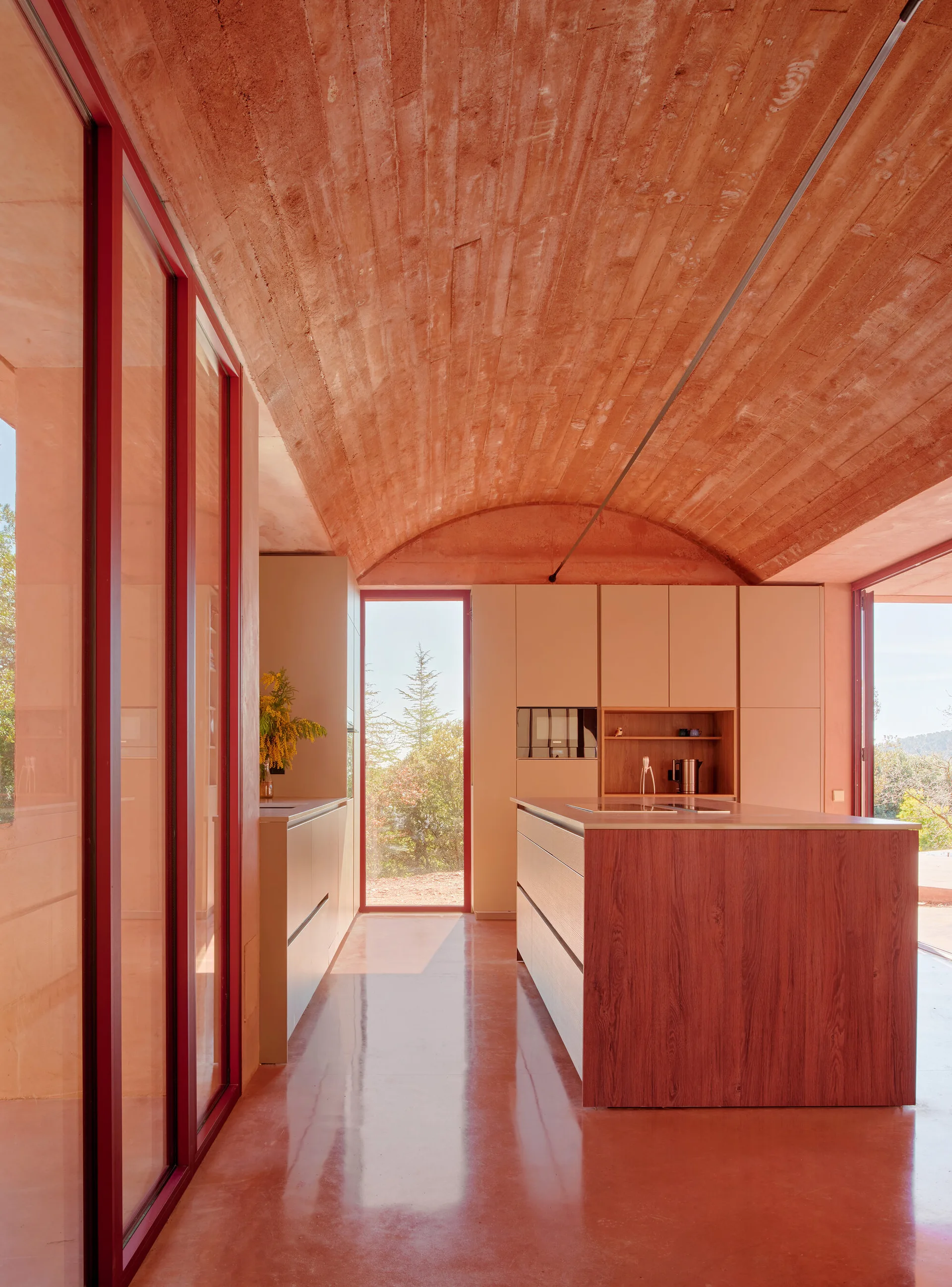 A minimalist kitchen features a vaulted terracotta concrete ceiling, matching polished floor, beige cabinetry, a wood island, and red-framed glass doors.