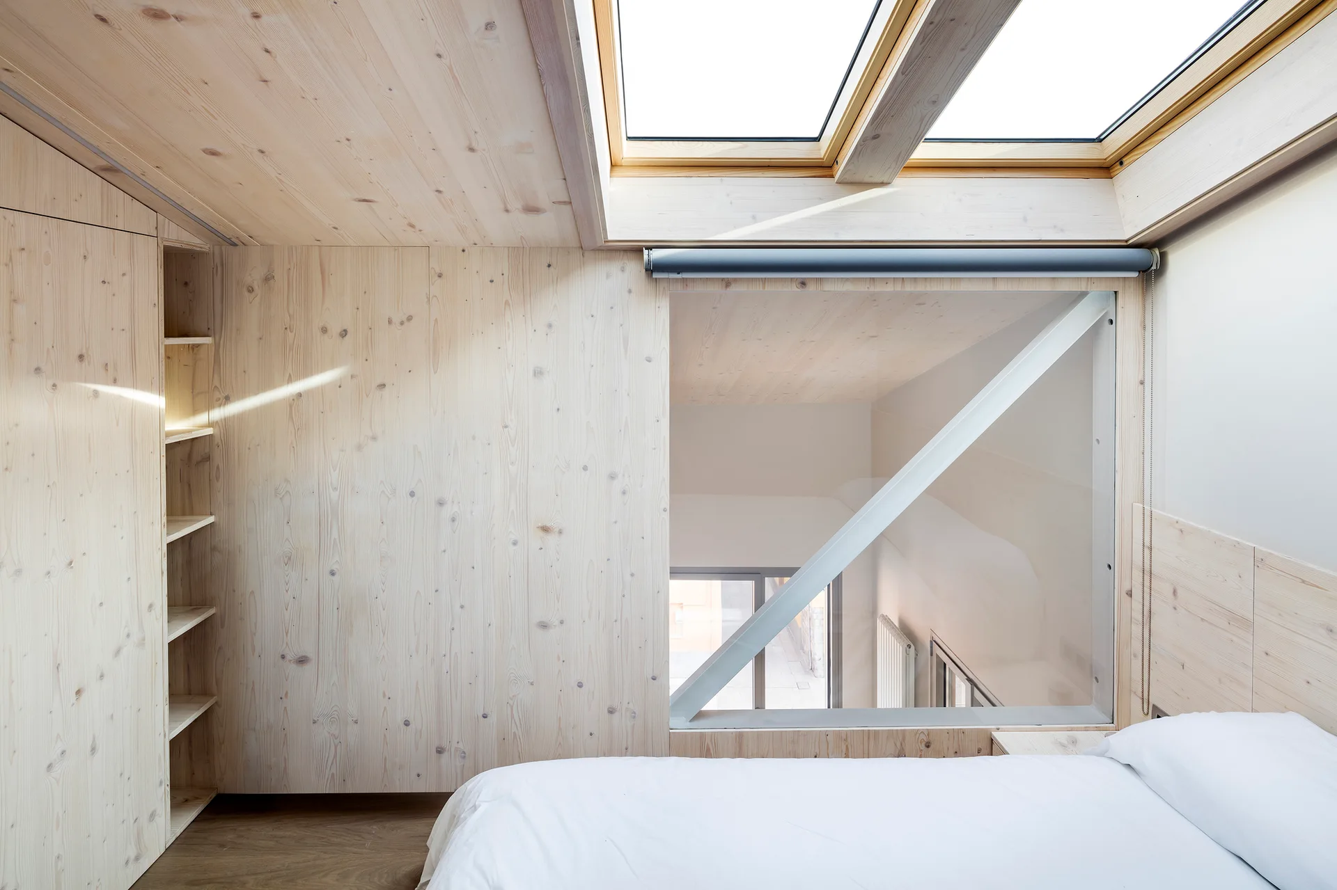 A minimalist attic bedroom features light wood paneling on the walls and ceiling, a large skylight, an interior window, and a bed with white sheets.