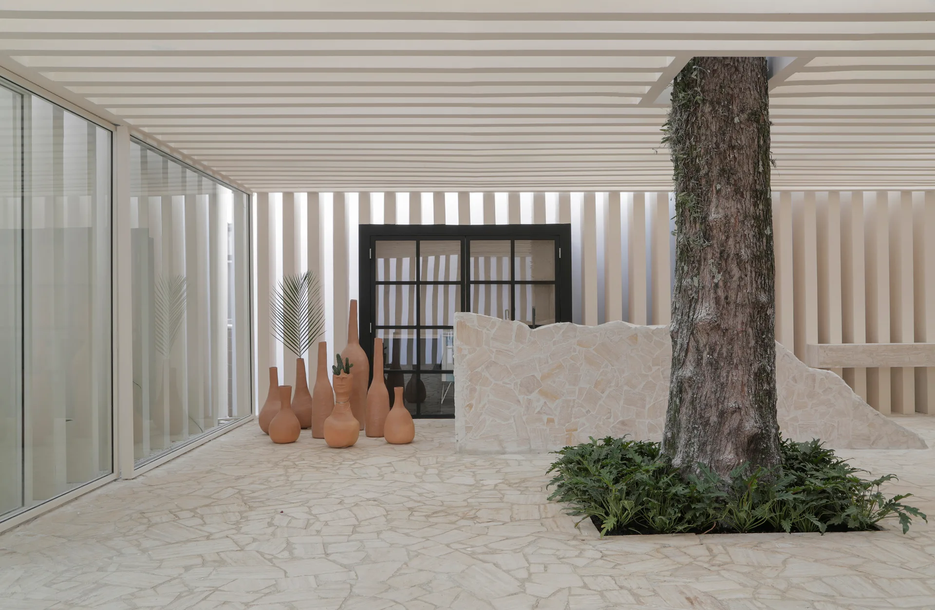 A modern covered patio features irregular flagstone paving, a slatted beige ceiling, a central tree, and a collection of tall terracotta pots.
